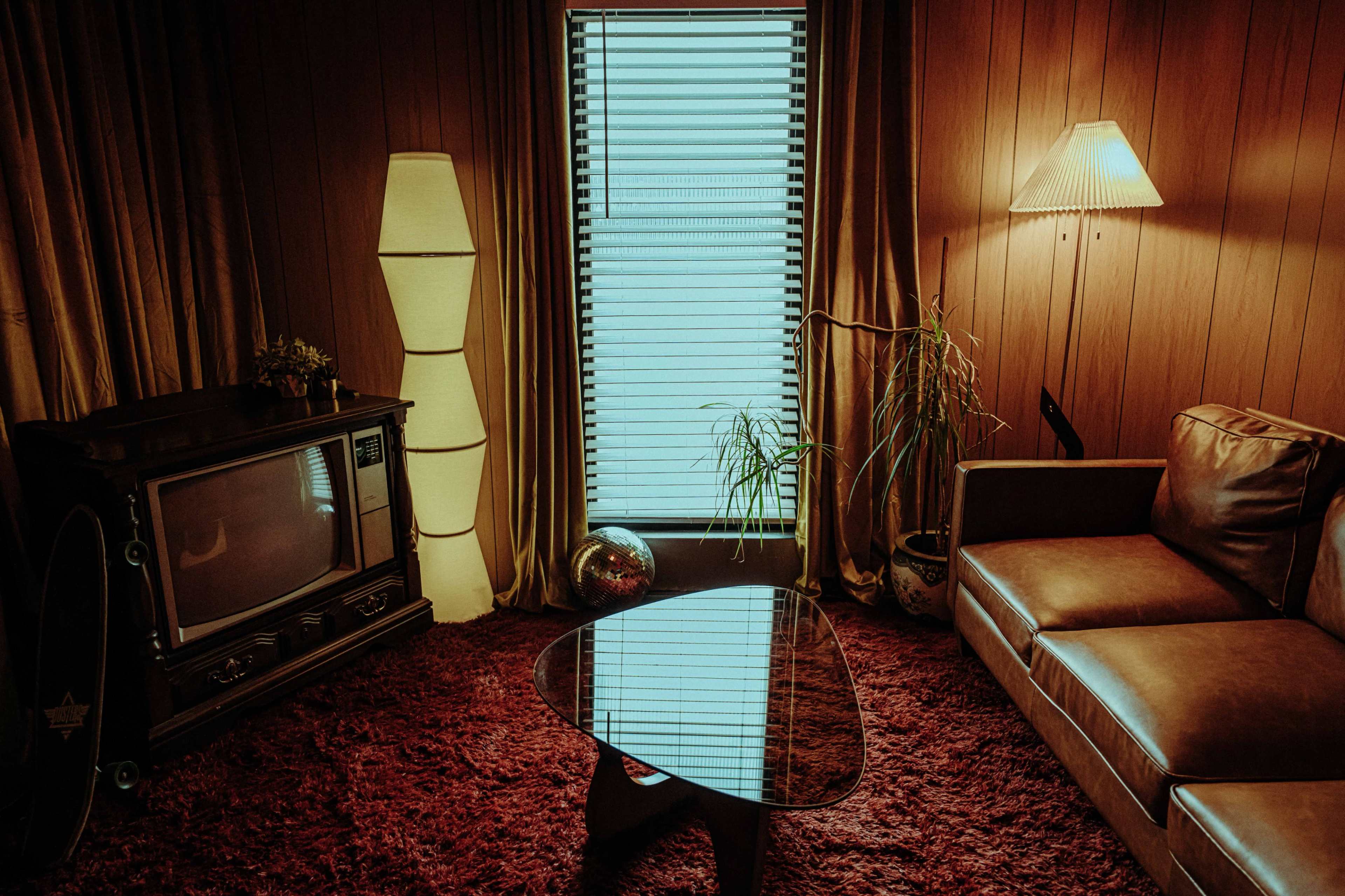 The image shows a retro-style living room with a brown leather sofa, a glass coffee table, a large television, and a window adorned with vertical blinds.