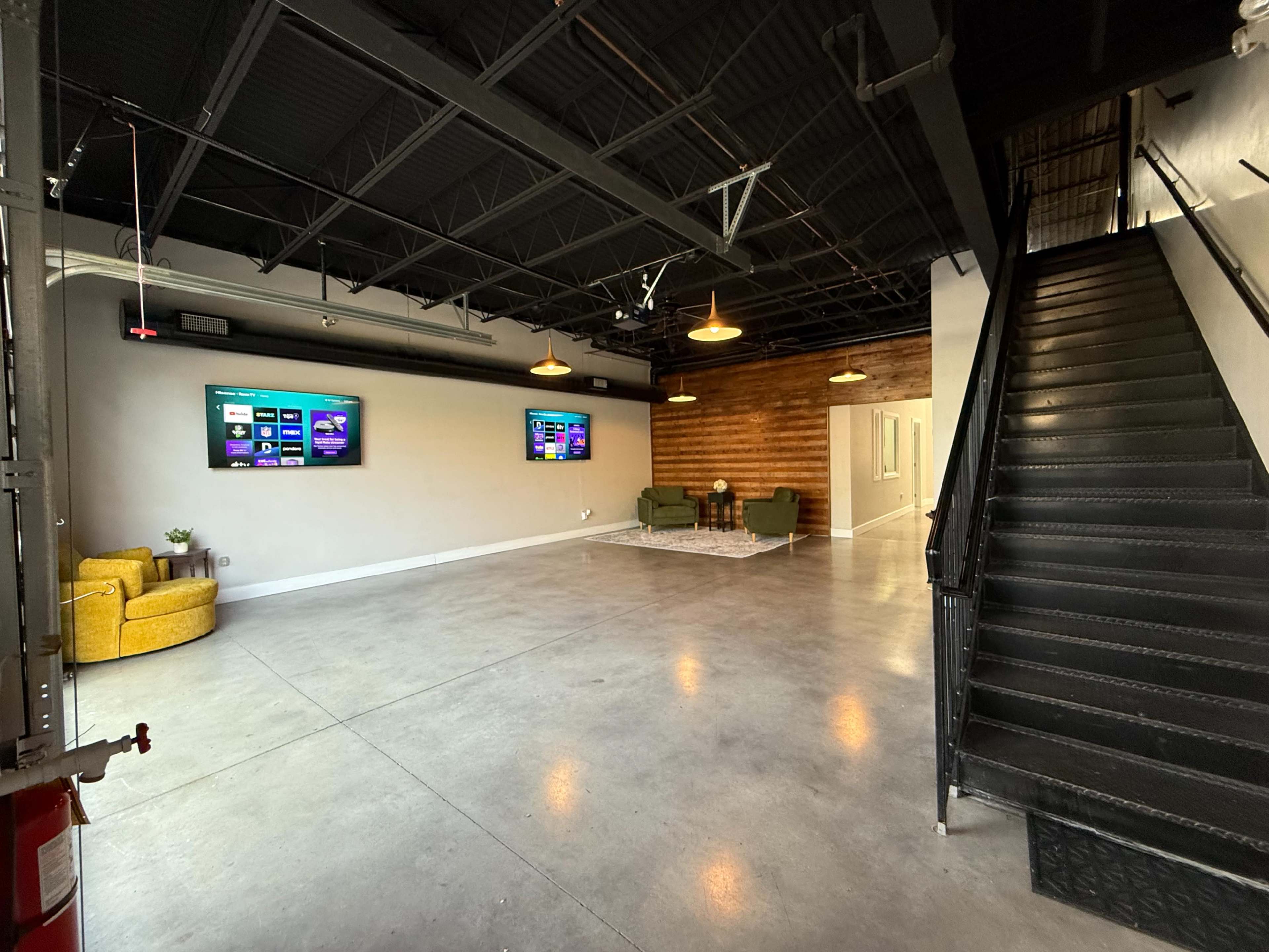 The image shows a modern, spacious interior with two television screens mounted on the wall, a yellow armchair, a green sofa, and a staircase leading to an upper level.