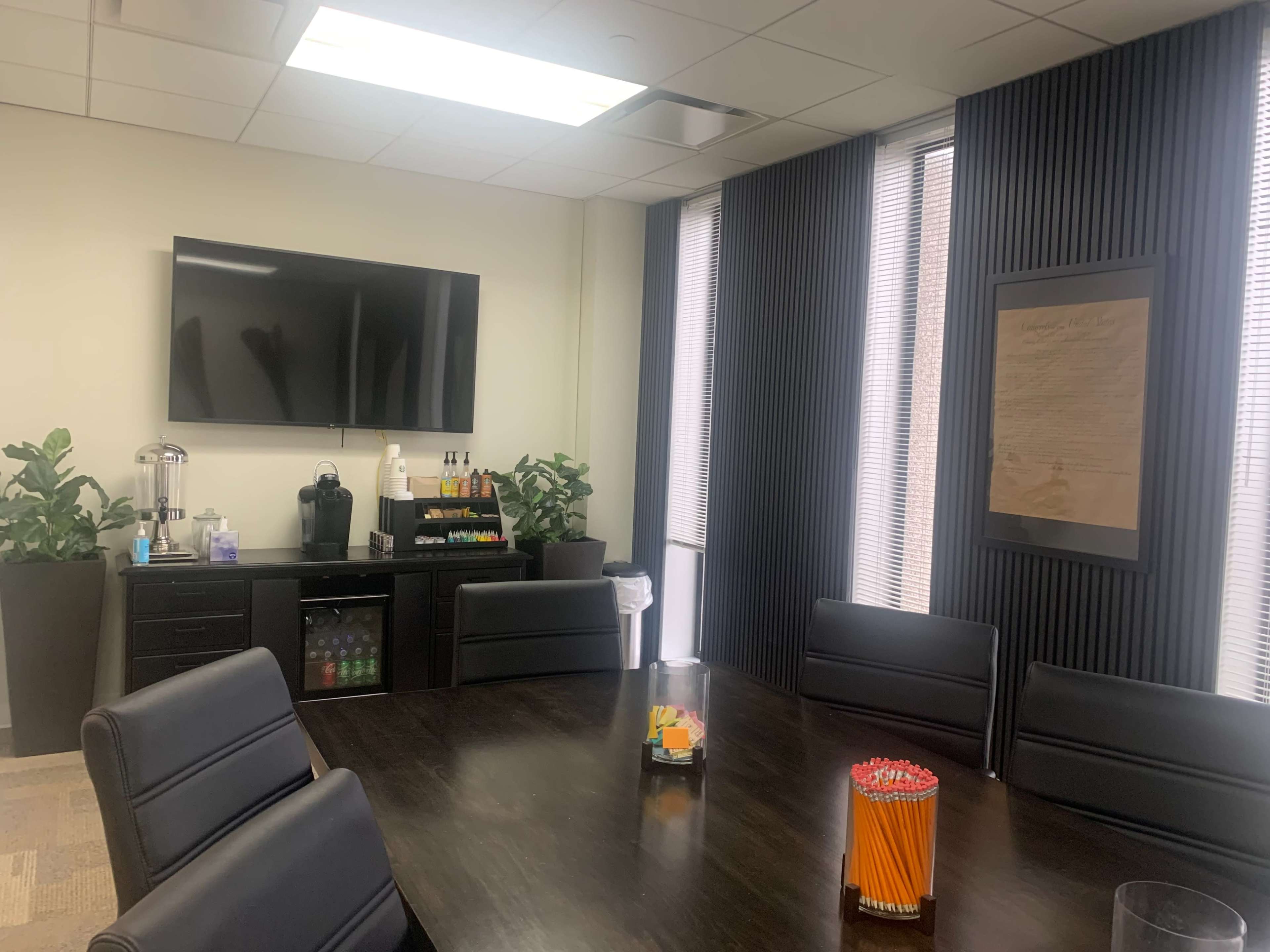 A conference room with a large table surrounded by black chairs, a television mounted on the wall, and a small refreshment area featuring plants, beverages, and a framed document.