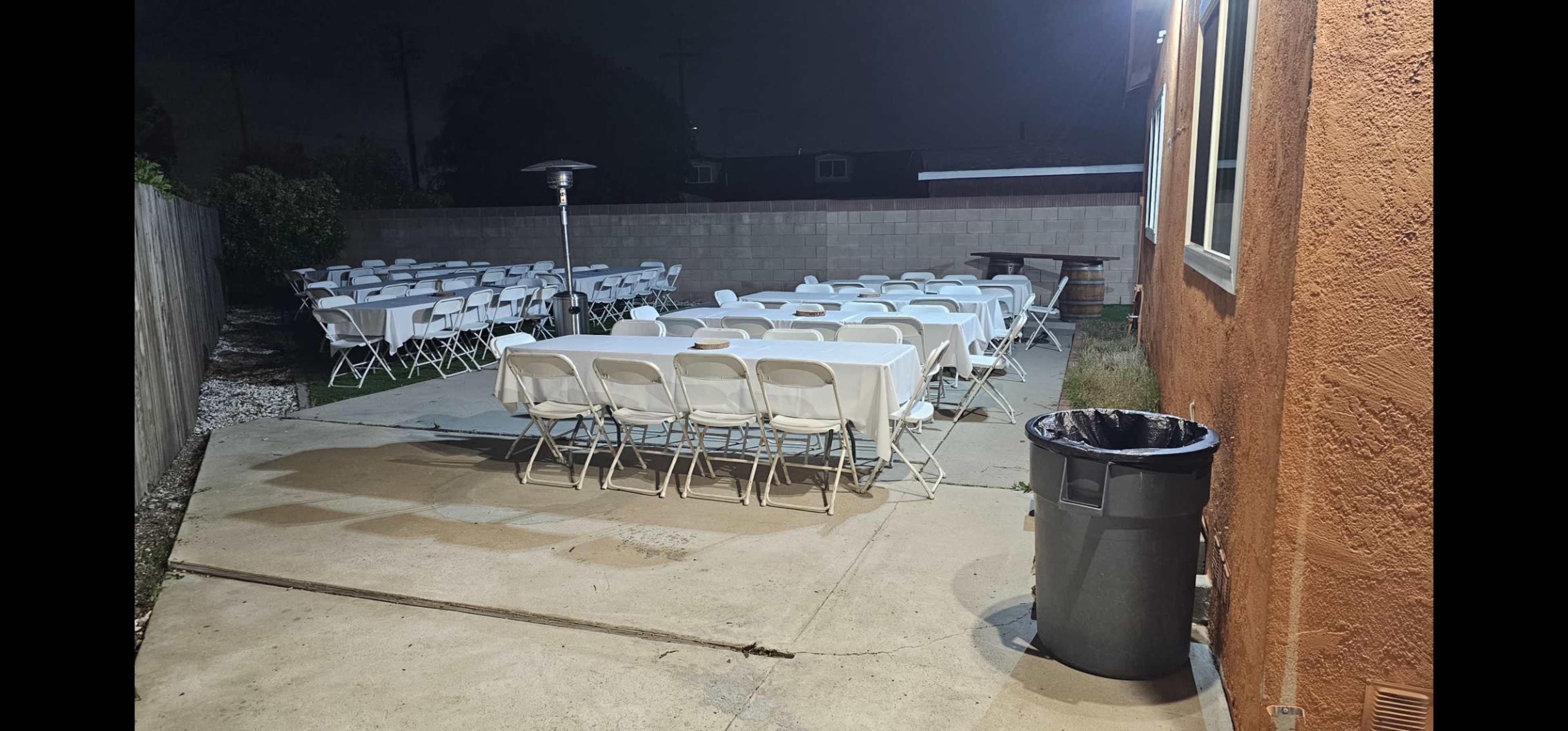 A row of white folding chairs is arranged around a patio area next to a wall, with a trash can in the corner and a heater in the background.