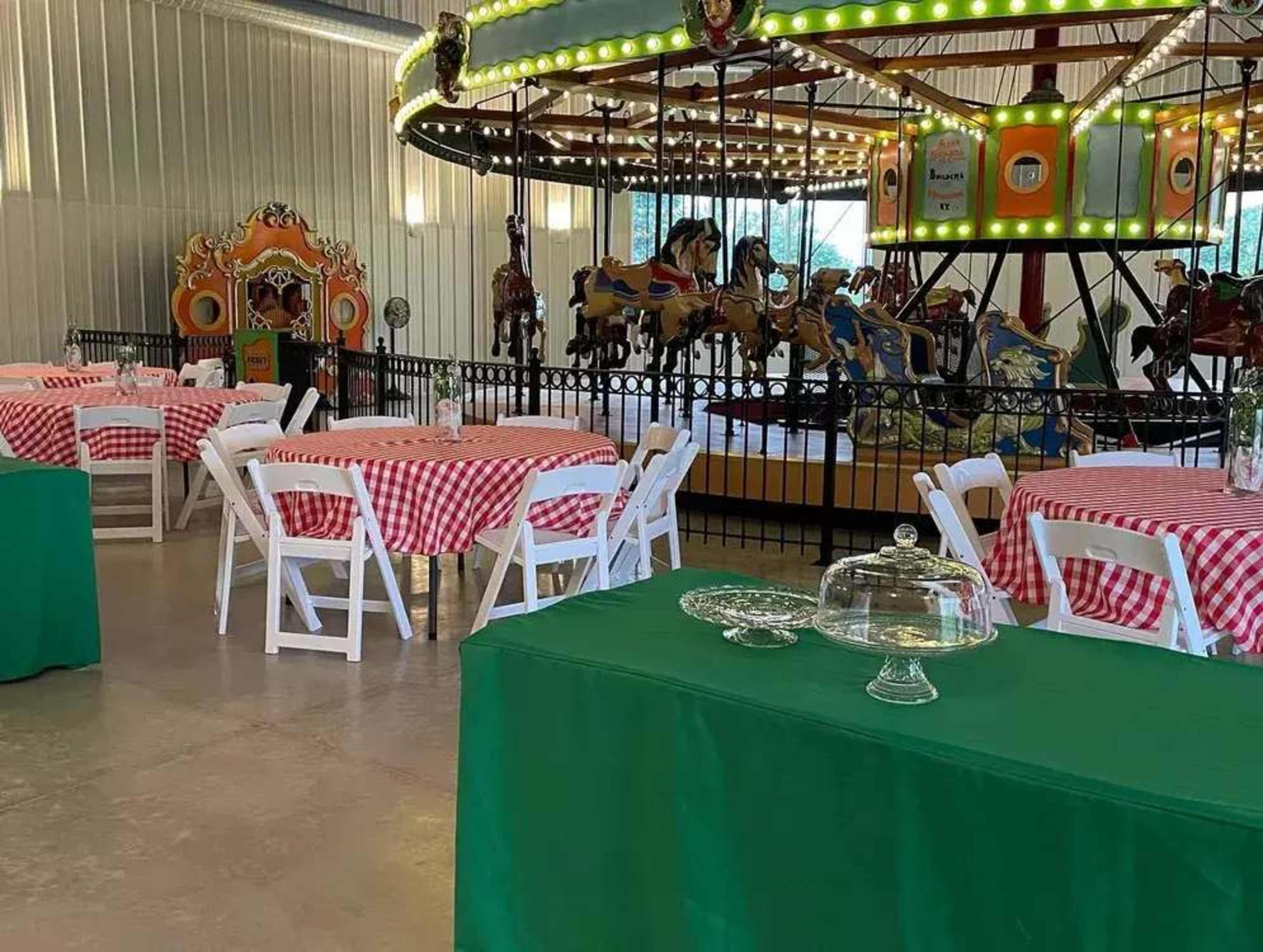The image shows a party venue with green tables covered by checkered tablecloths, a carousel with decorative horses in the background, and lighting around the carousel.