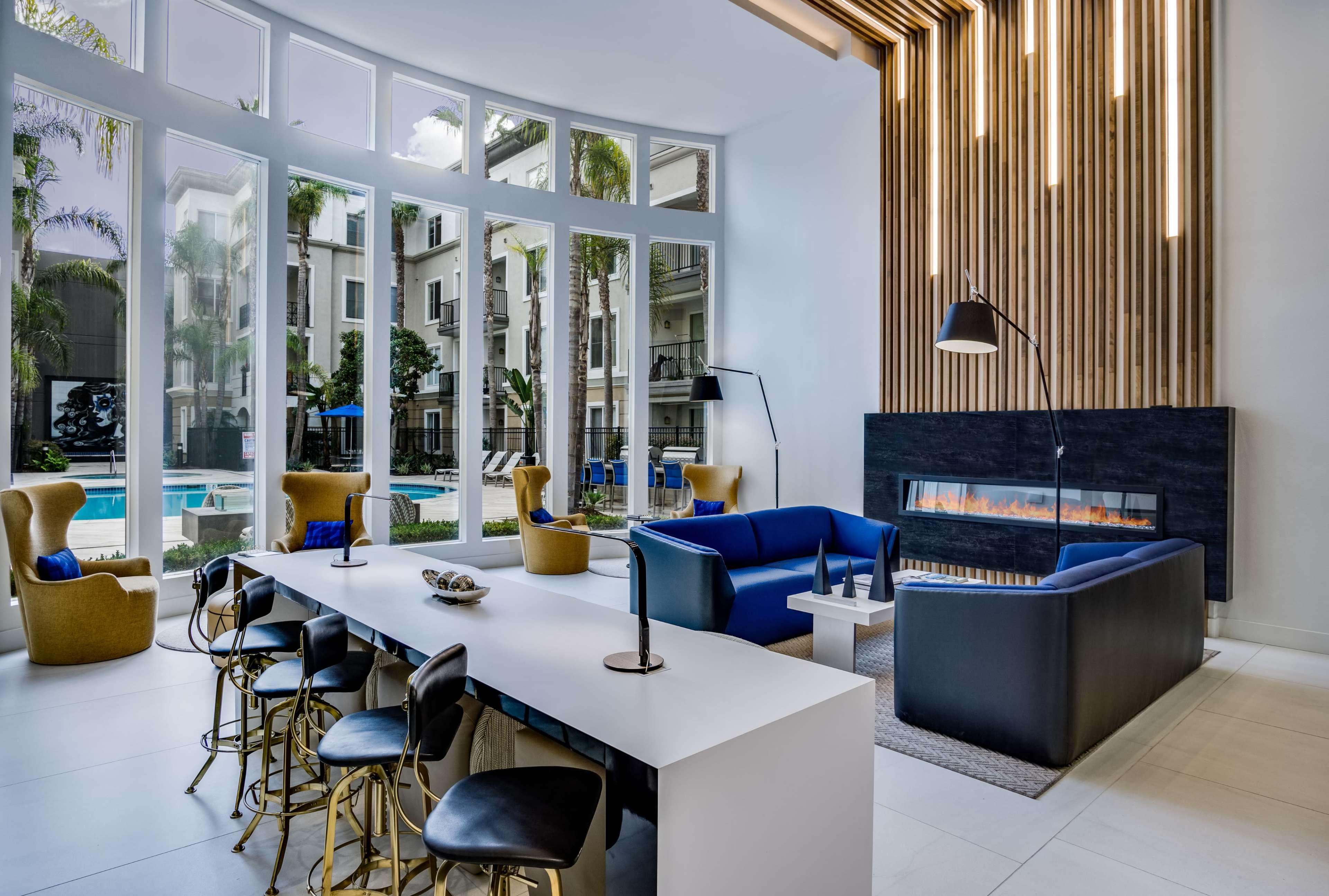 A modern indoor lounge area with a large table, stylish seating, and a fireplace, overlooking a pool surrounded by palm trees.