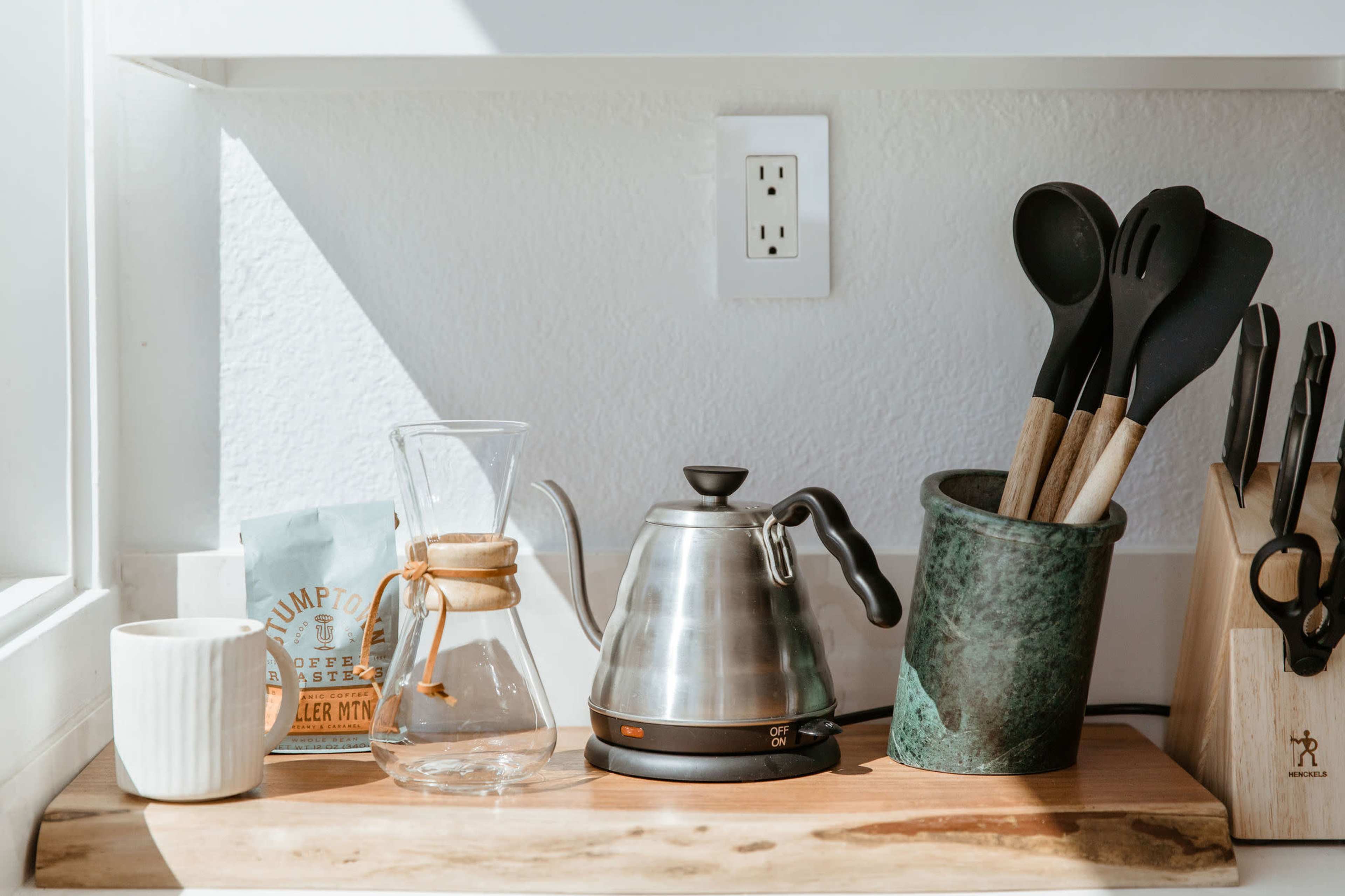 A clean kitchen countertop features a coffee setup with a kettle, glass carafe, a bag of coffee, a ceramic mug, and cooking utensils in a holder.