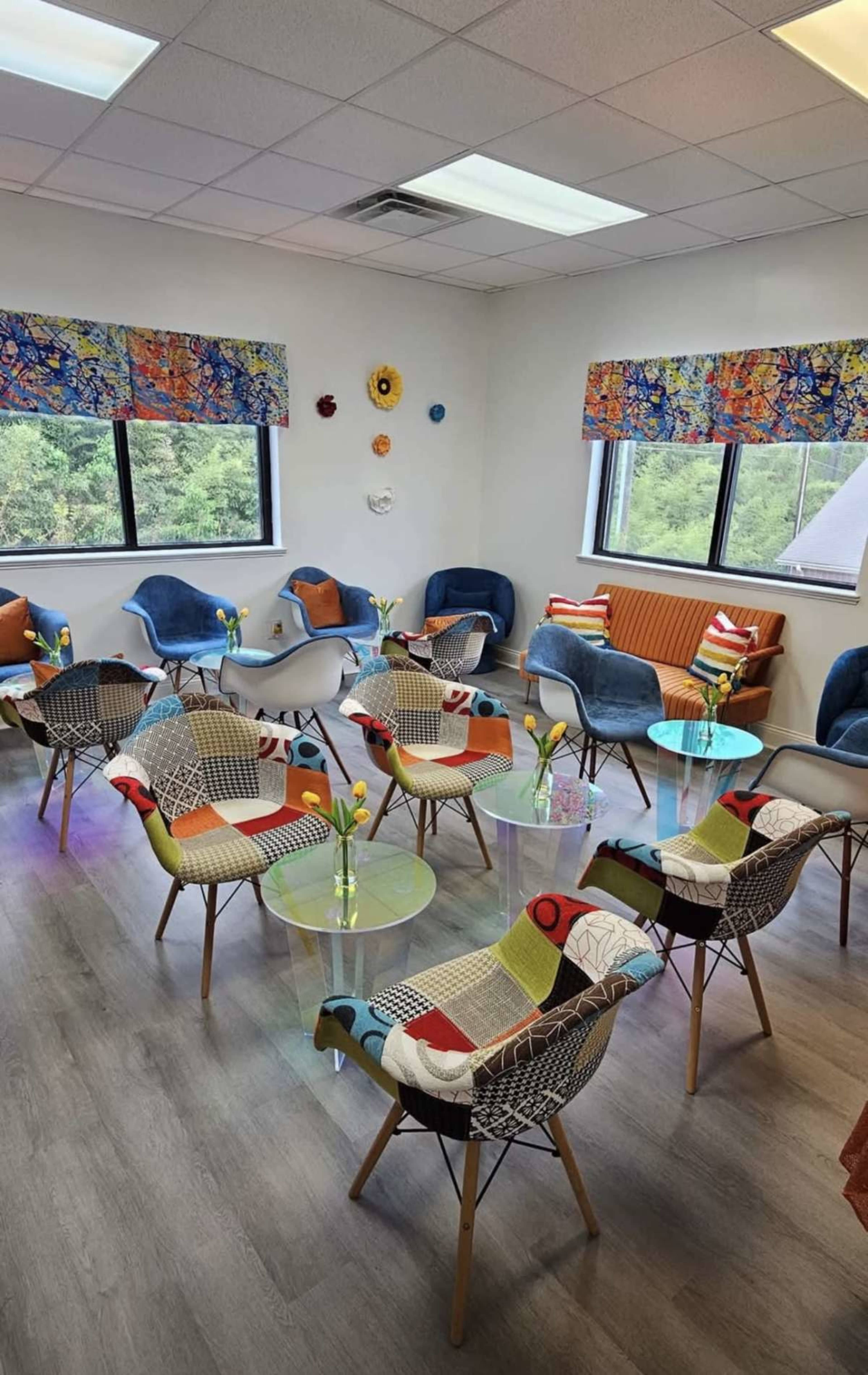The image shows a brightly lit room with multiple colorful chairs and tables arranged in a seating area, accompanied by patterned window treatments and decorative elements on the walls.