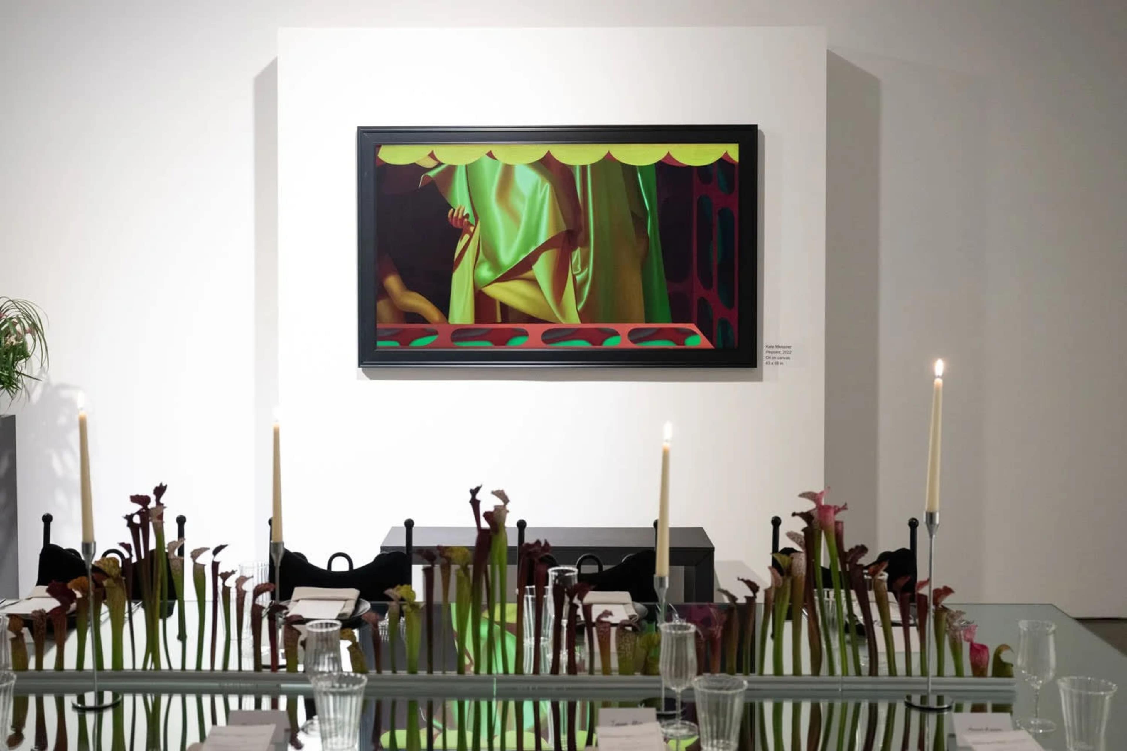 The image shows a modern art exhibition featuring a colorful painting displayed on a white wall, with an elegantly arranged table in the foreground adorned with unique floral centerpieces and candles.