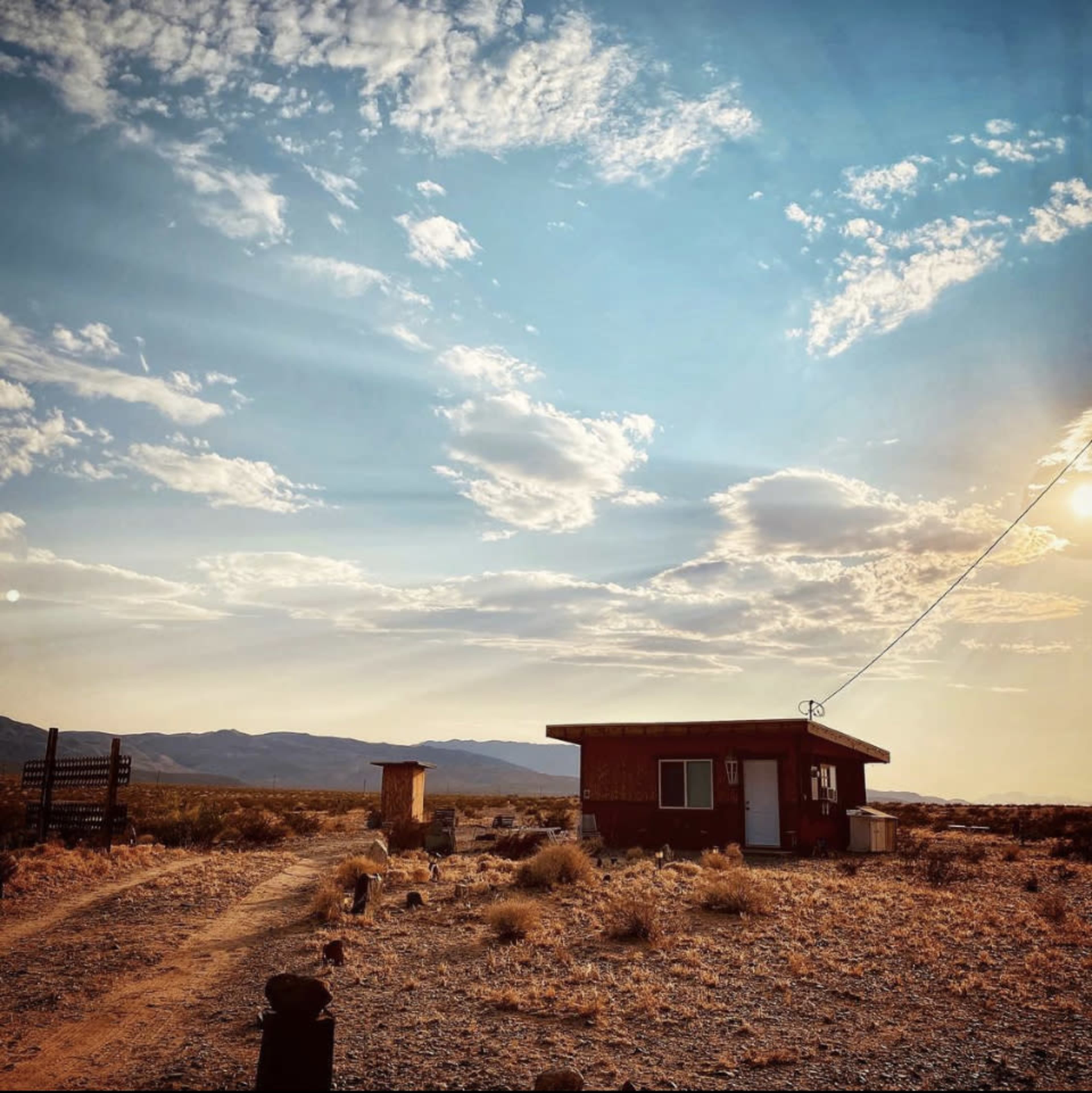 A small wooden building sits along a dirt path in a vast, arid landscape under a blue sky with scattered clouds.