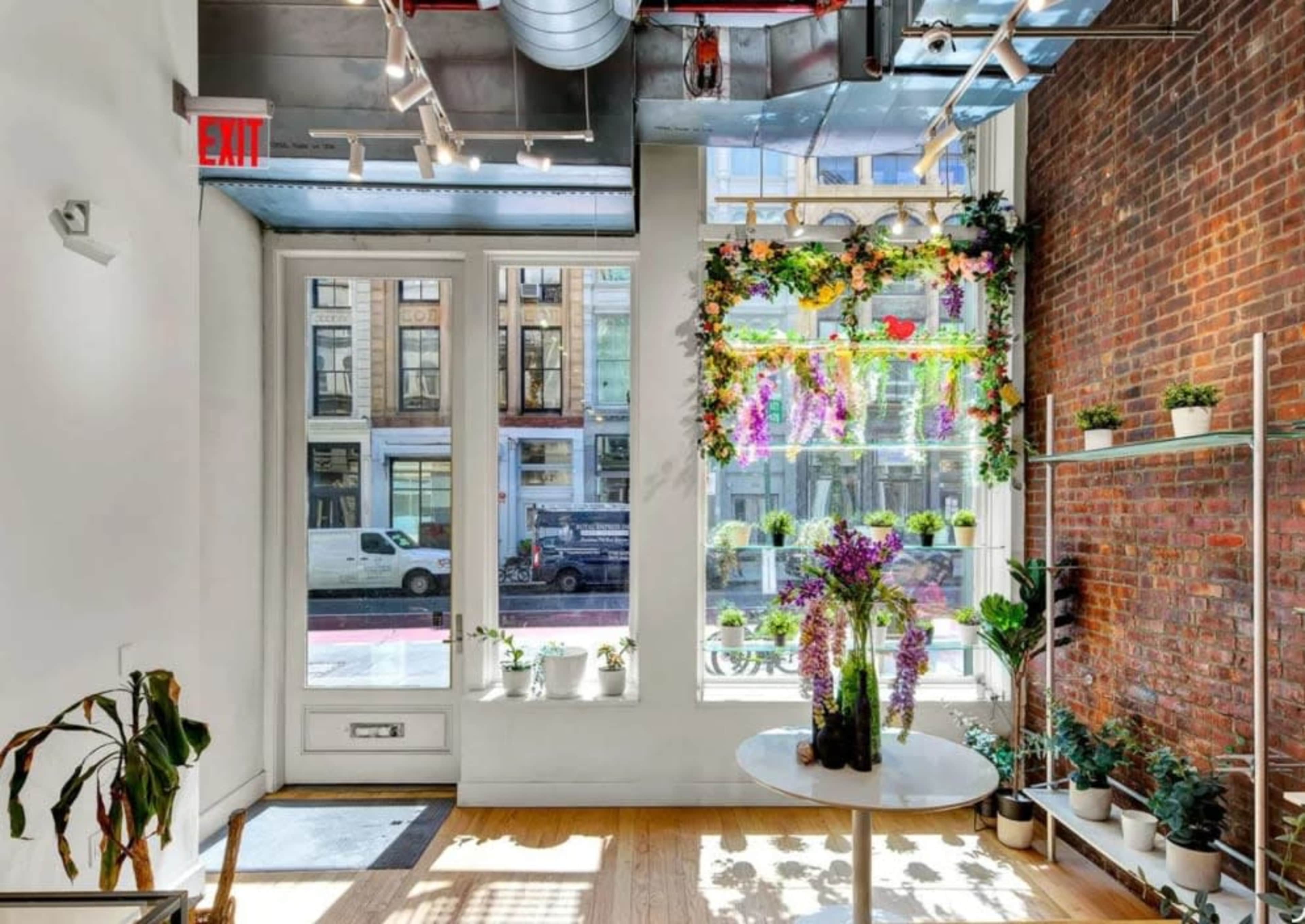 The interior of a bright room features large windows adorned with floral decorations, a round table in the center, and plants on shelves along the brick wall.