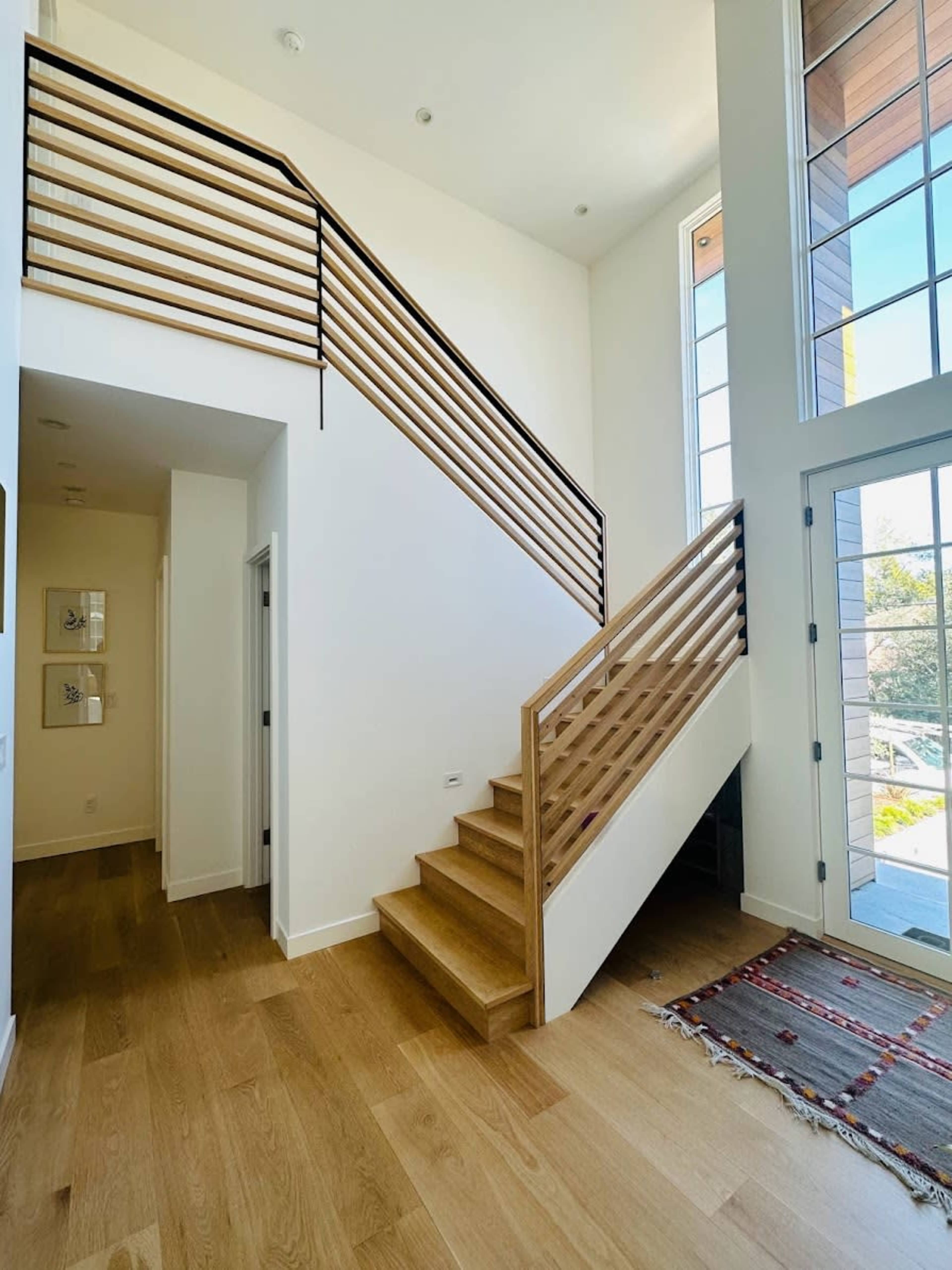 A modern entrance hall with a wooden staircase, large windows, and a woven rug on the hardwood floor.