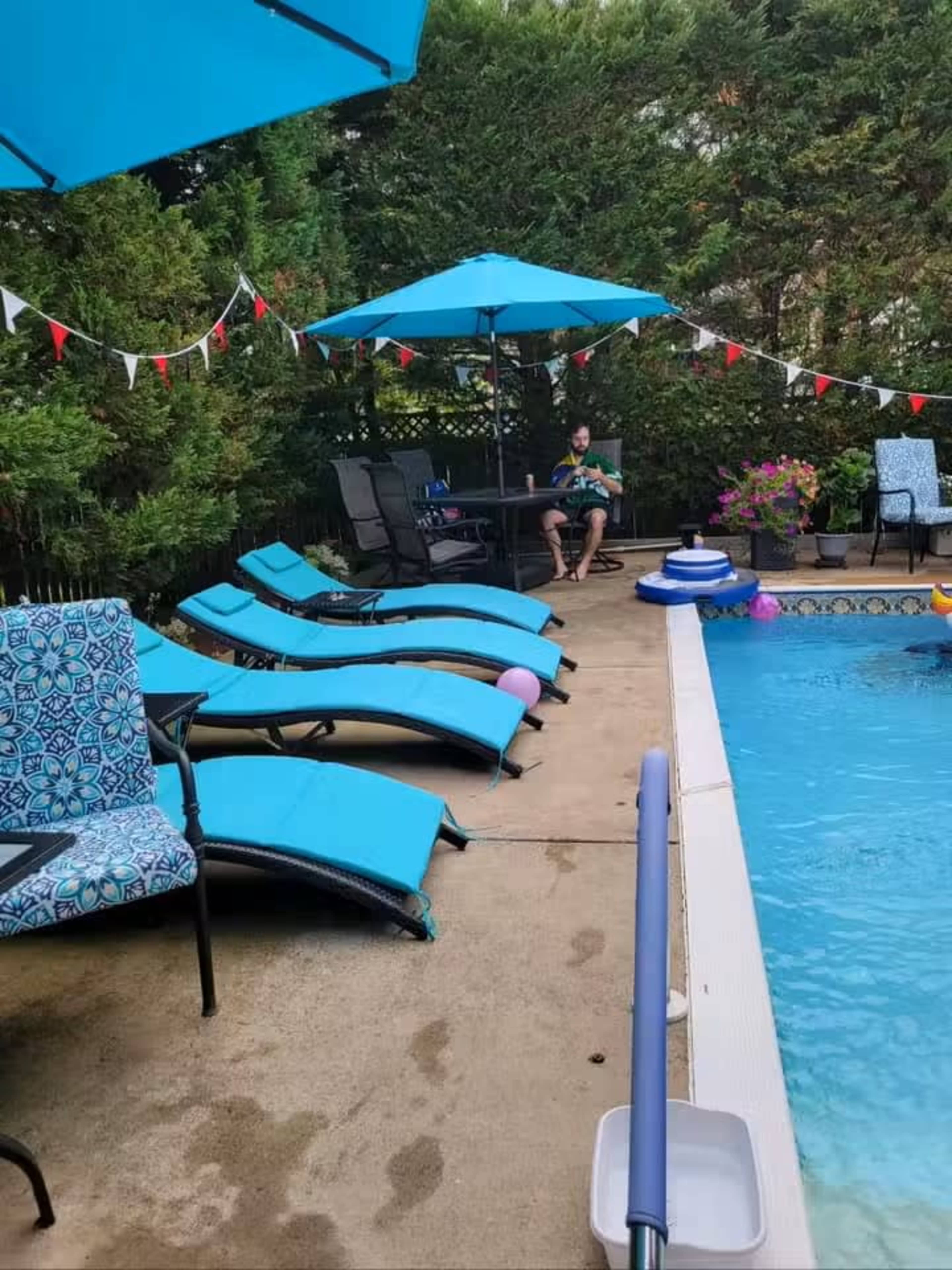 A patio area with blue loungers and umbrellas surrounds a swimming pool where a person is sitting at a table.