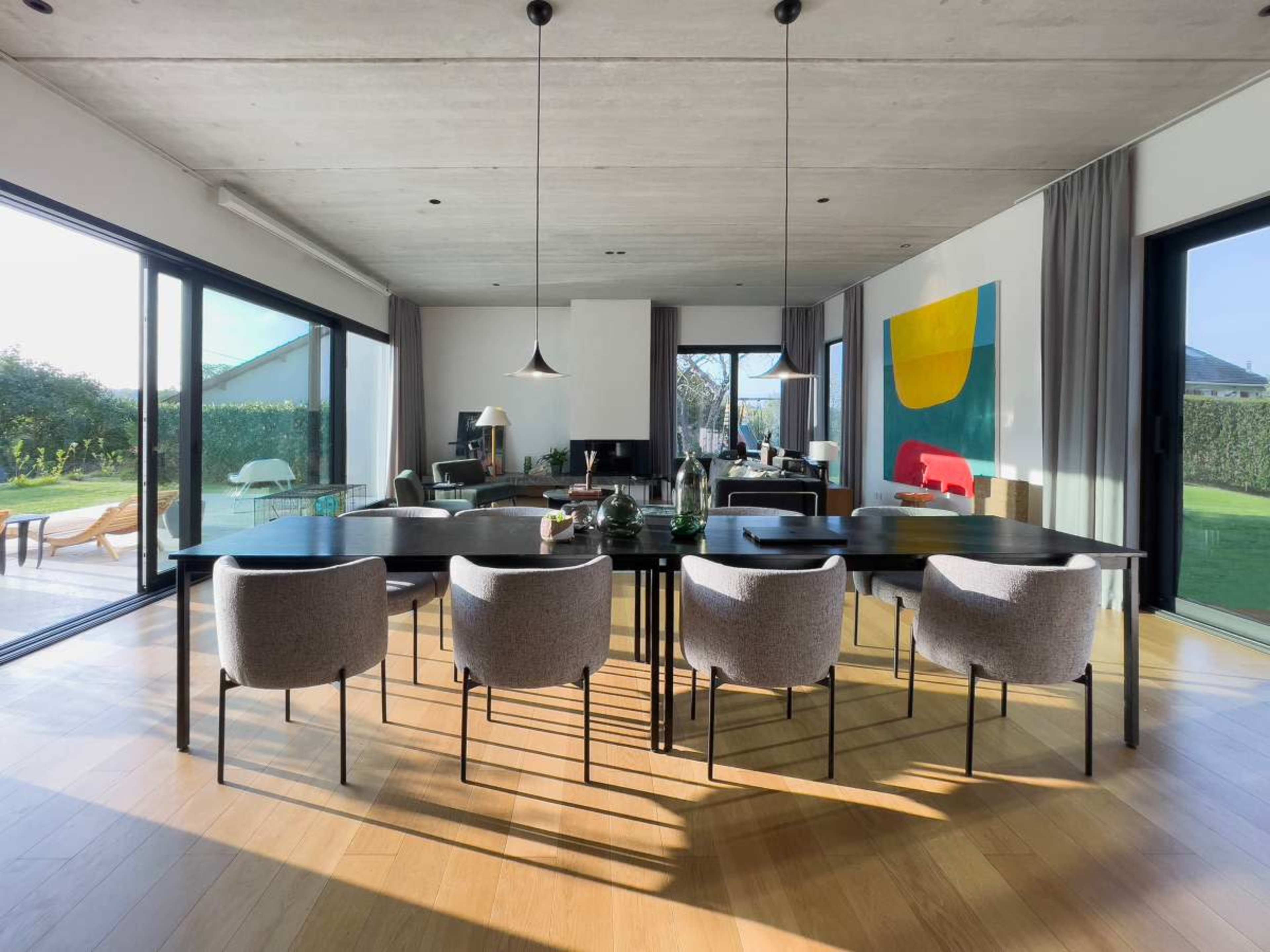 A modern dining area features a large table surrounded by upholstered chairs, with large glass doors opening to a garden.