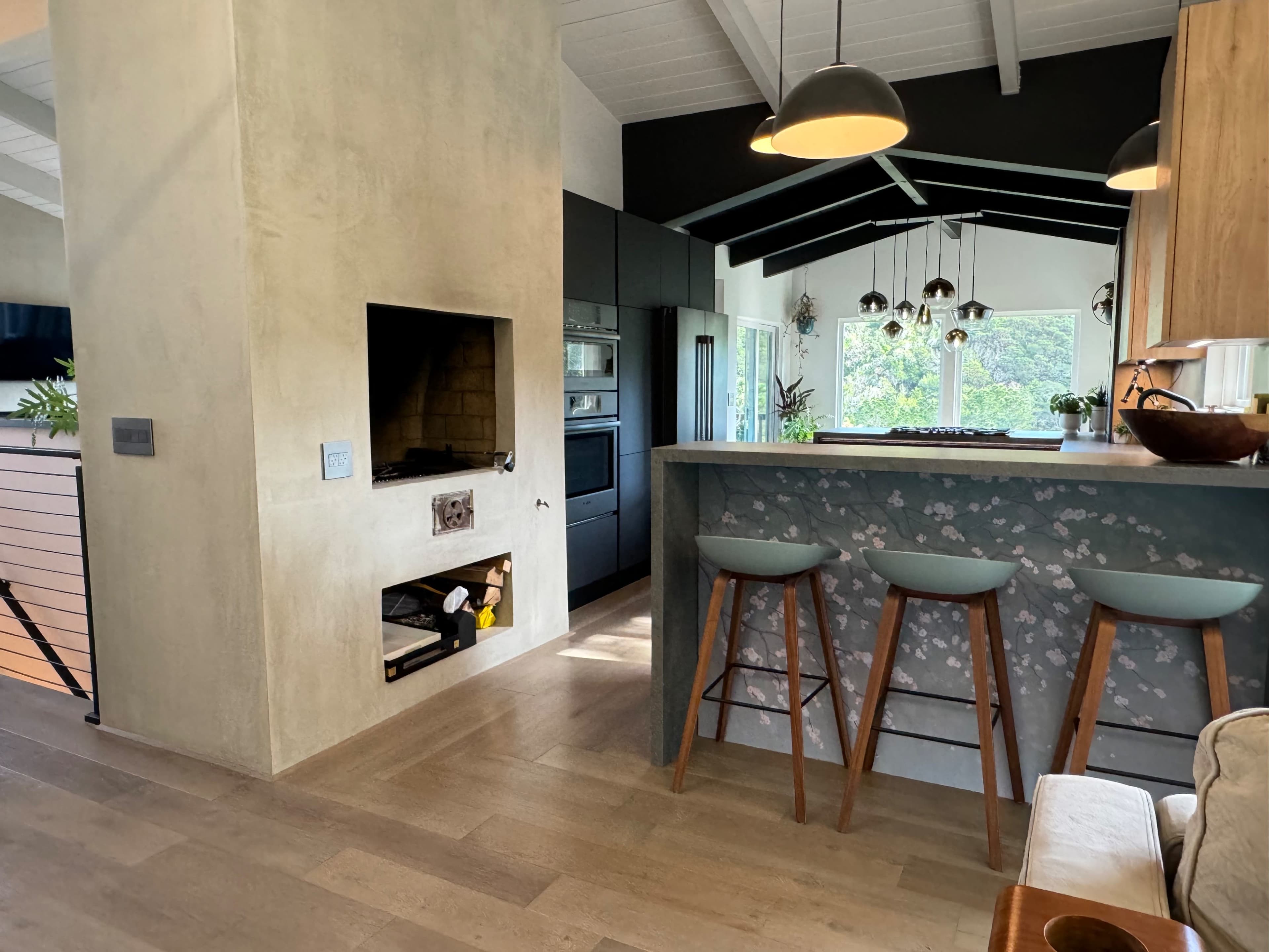 A modern kitchen with a bar area featuring three stools, a built-in fireplace, and large windows allowing natural light to enter.