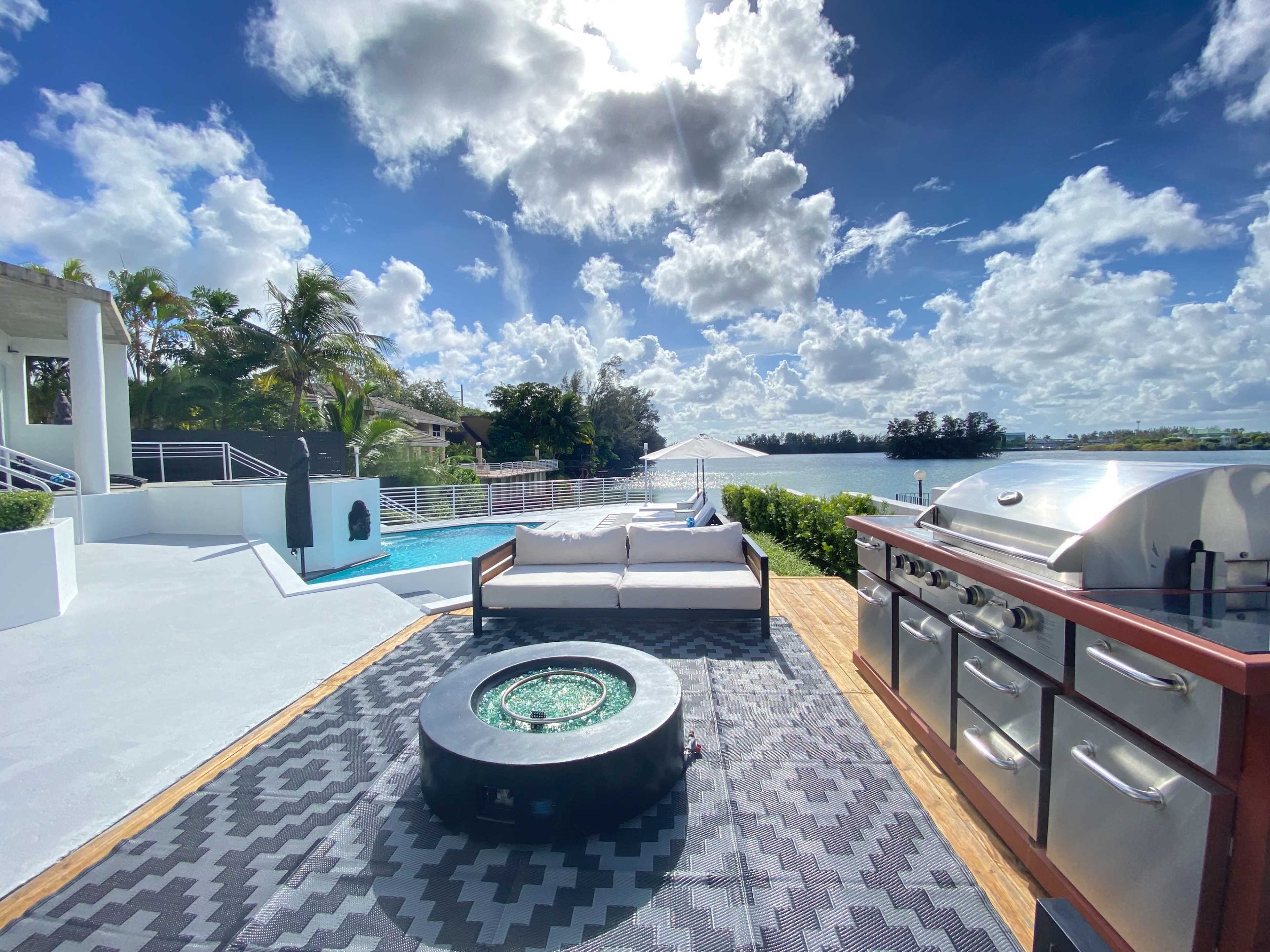 The scene shows a backyard patio featuring a grill, a circular fire pit, and a seating area overlooking a swimming pool and a body of water under a cloudy sky.