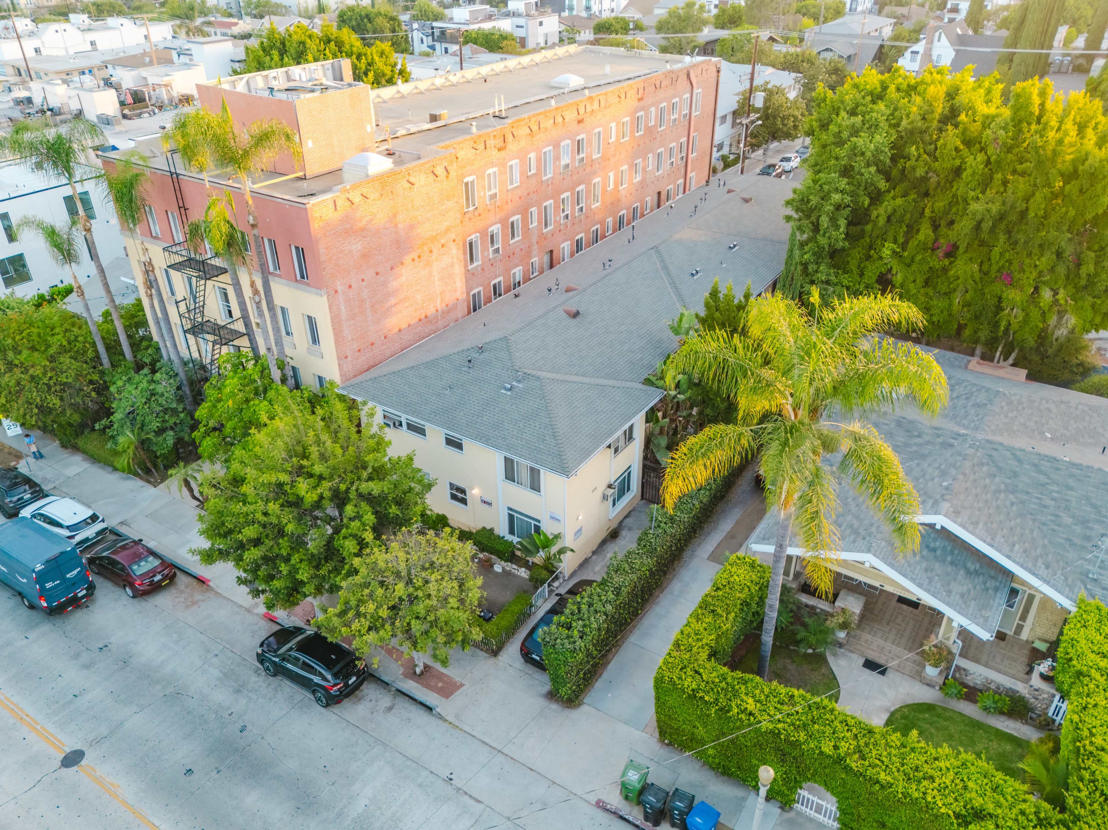 The image shows an aerial view of a neighborhood featuring a mix of low-rise buildings, palm trees, and parked cars along a street.
