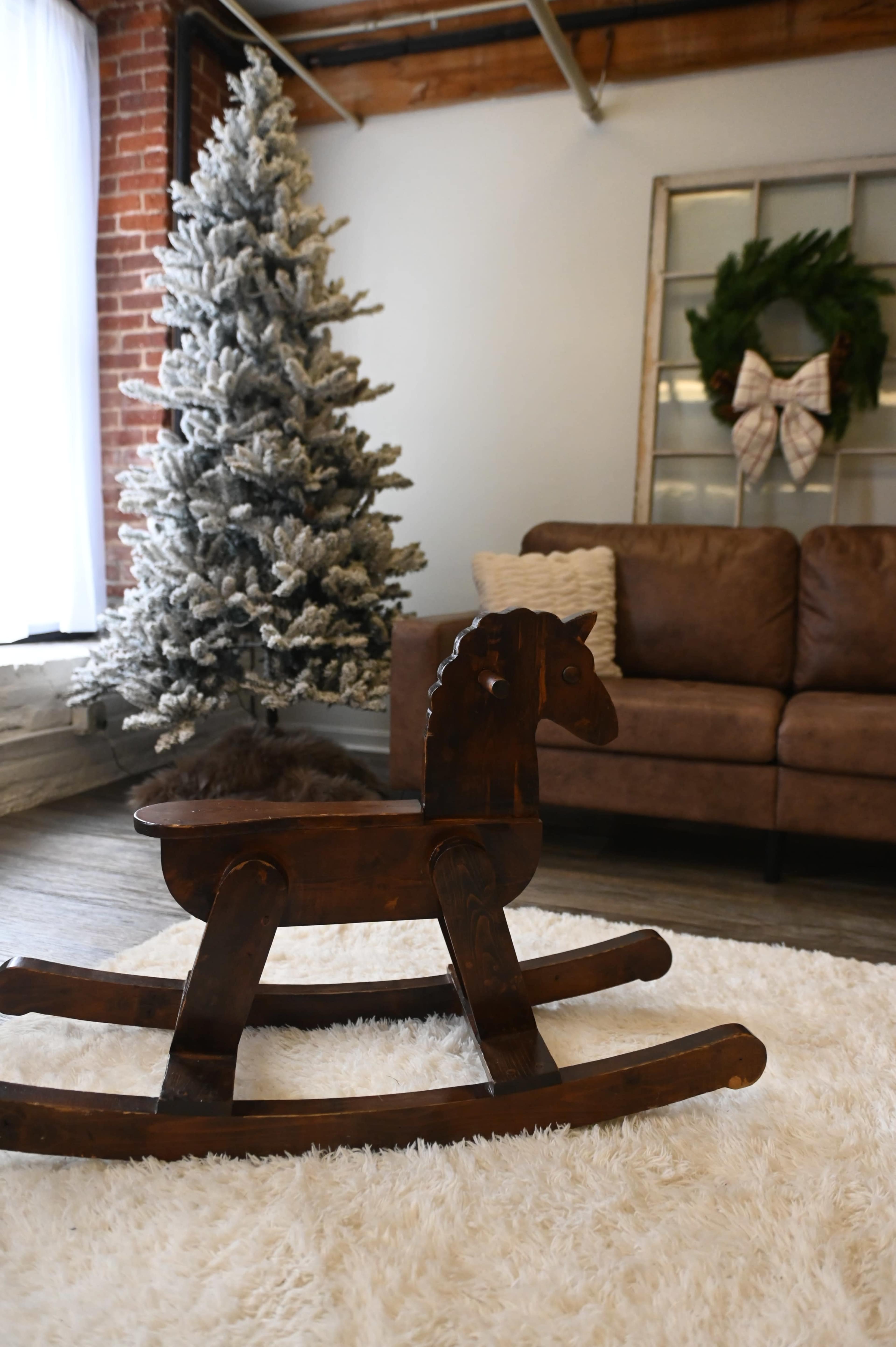 A wooden rocking horse sits on a plush rug in front of a Christmas tree and a brown leather sofa.