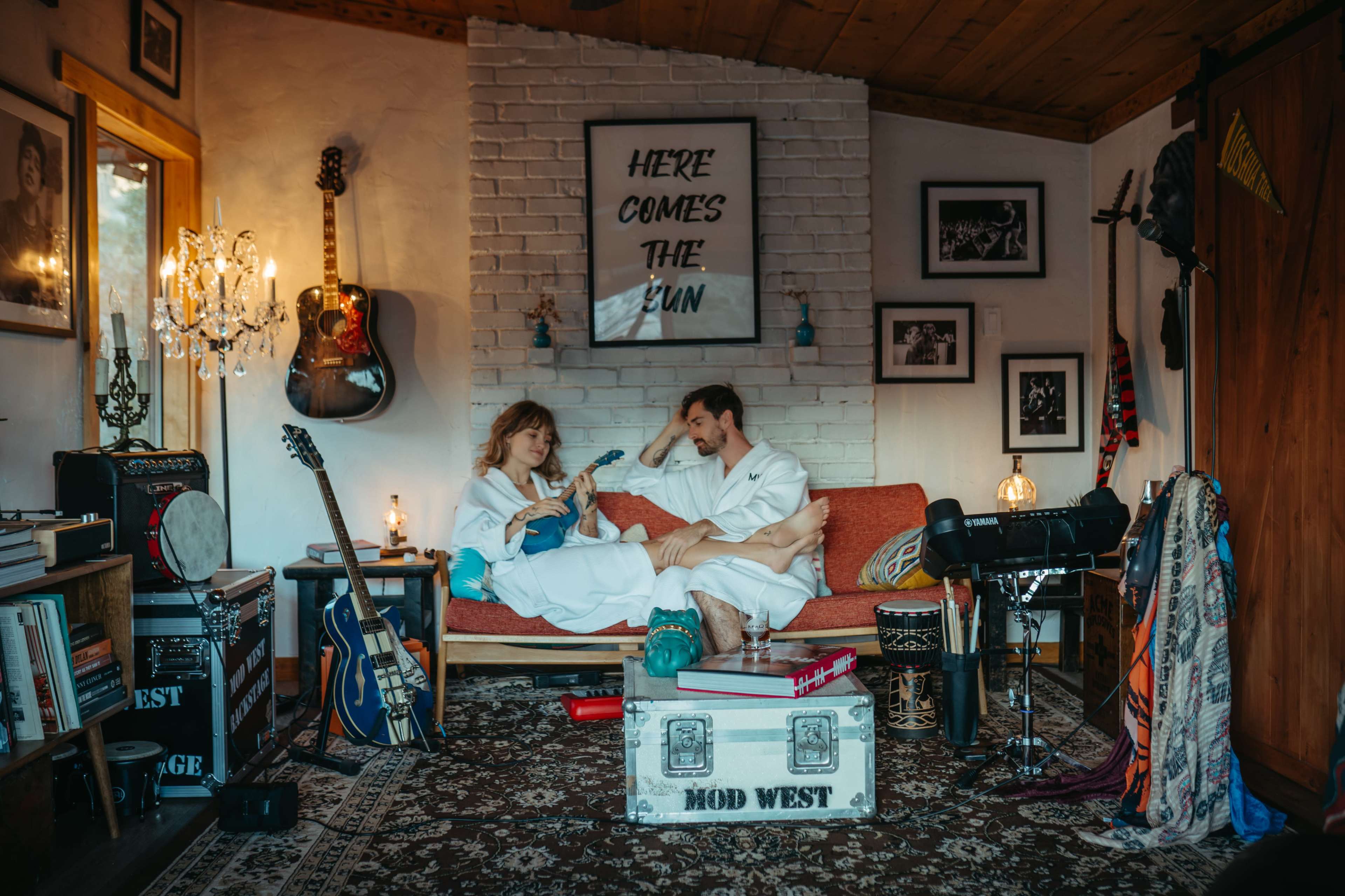 A man and a woman in bathrobes sit on a sofa in a cozy room decorated with musical instruments and photographs.