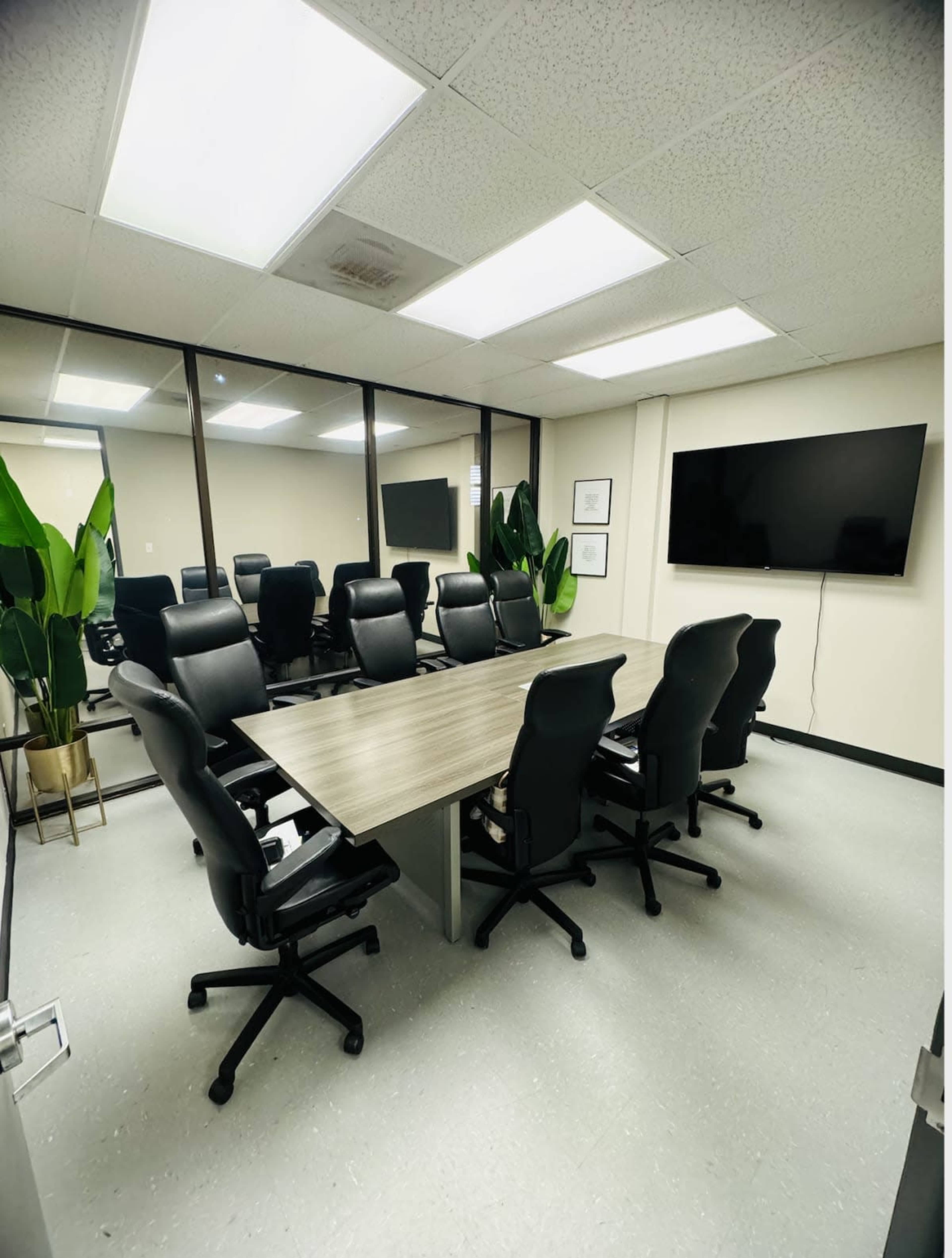 A modern conference room with a long table surrounded by black office chairs, and large windows facing a wall-mounted television.