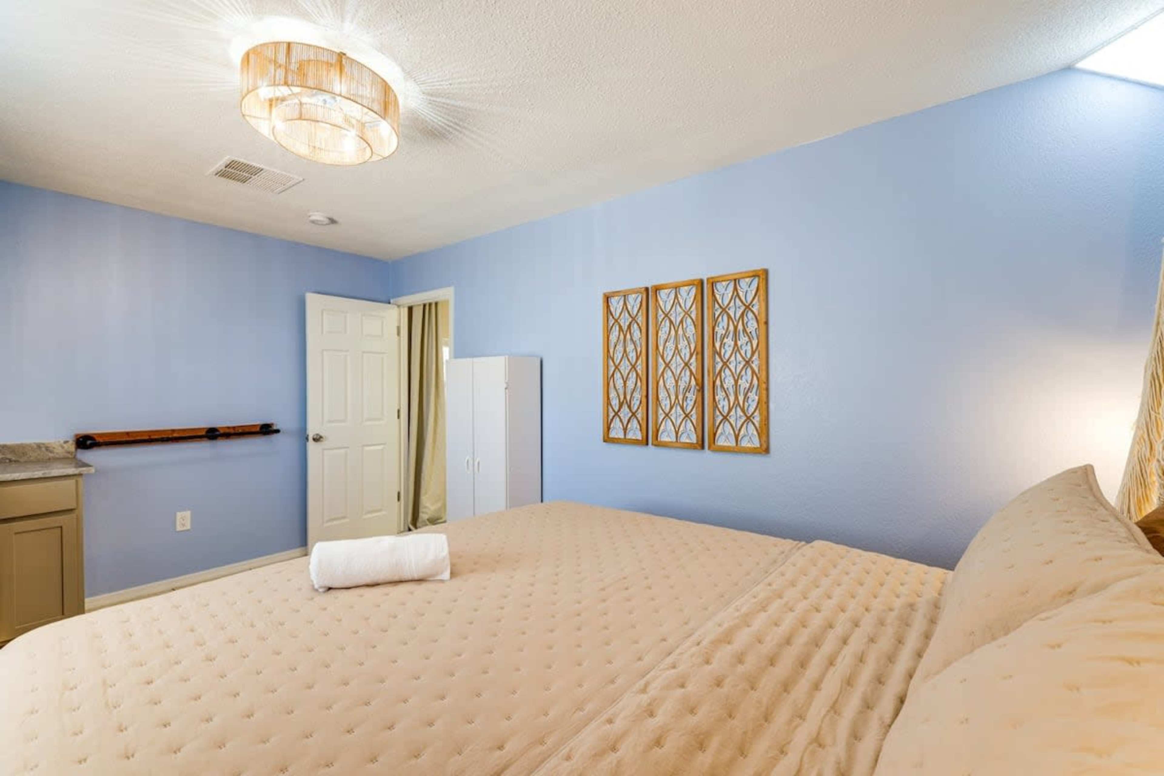The image shows a well-lit bedroom with a large bed, light blue walls, and framed decorations on the wall above a wardrobe.