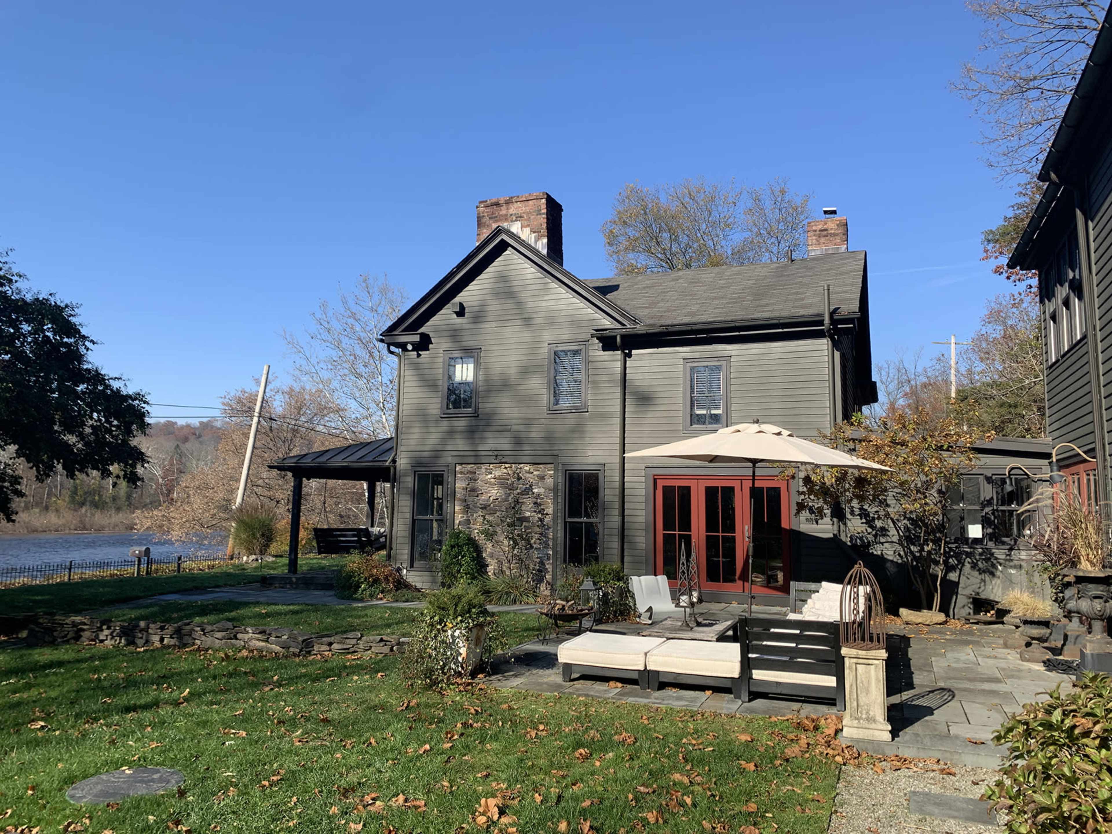A two-story gray house with a patio and outdoor seating overlooks a river, surrounded by trees and a lawn.