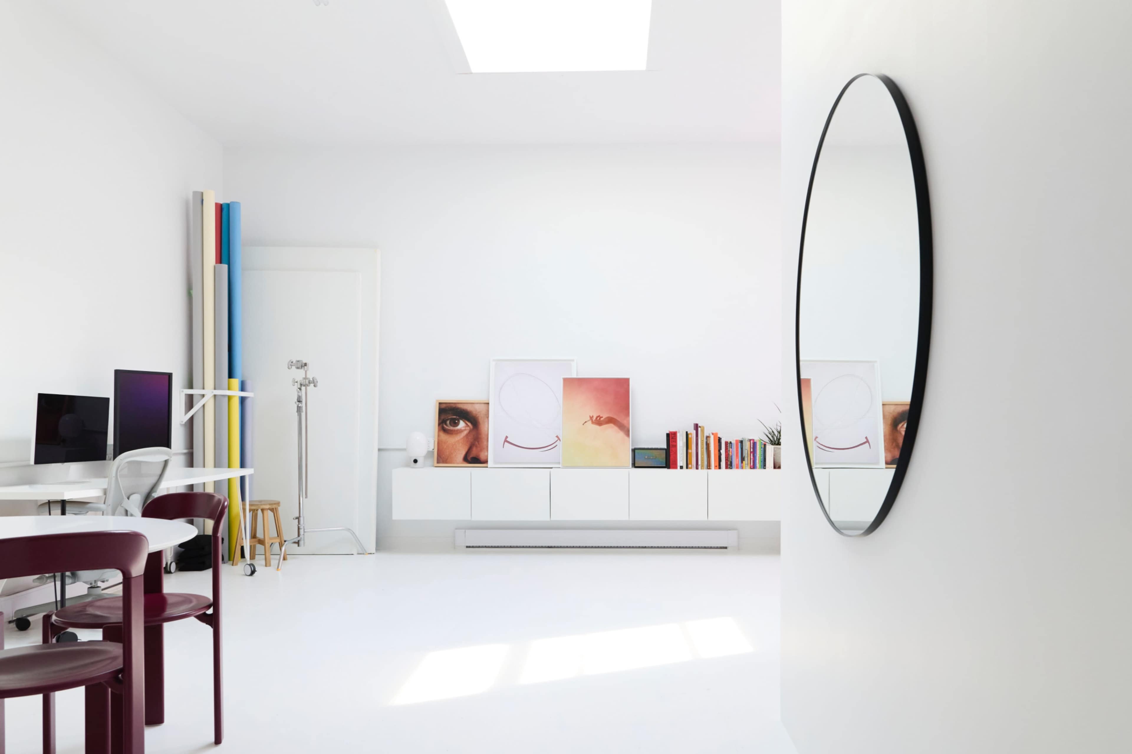 The image shows a bright, minimalist studio space featuring a circular mirror, white walls, several pieces of artwork, bookshelves, and furniture arranged neatly.