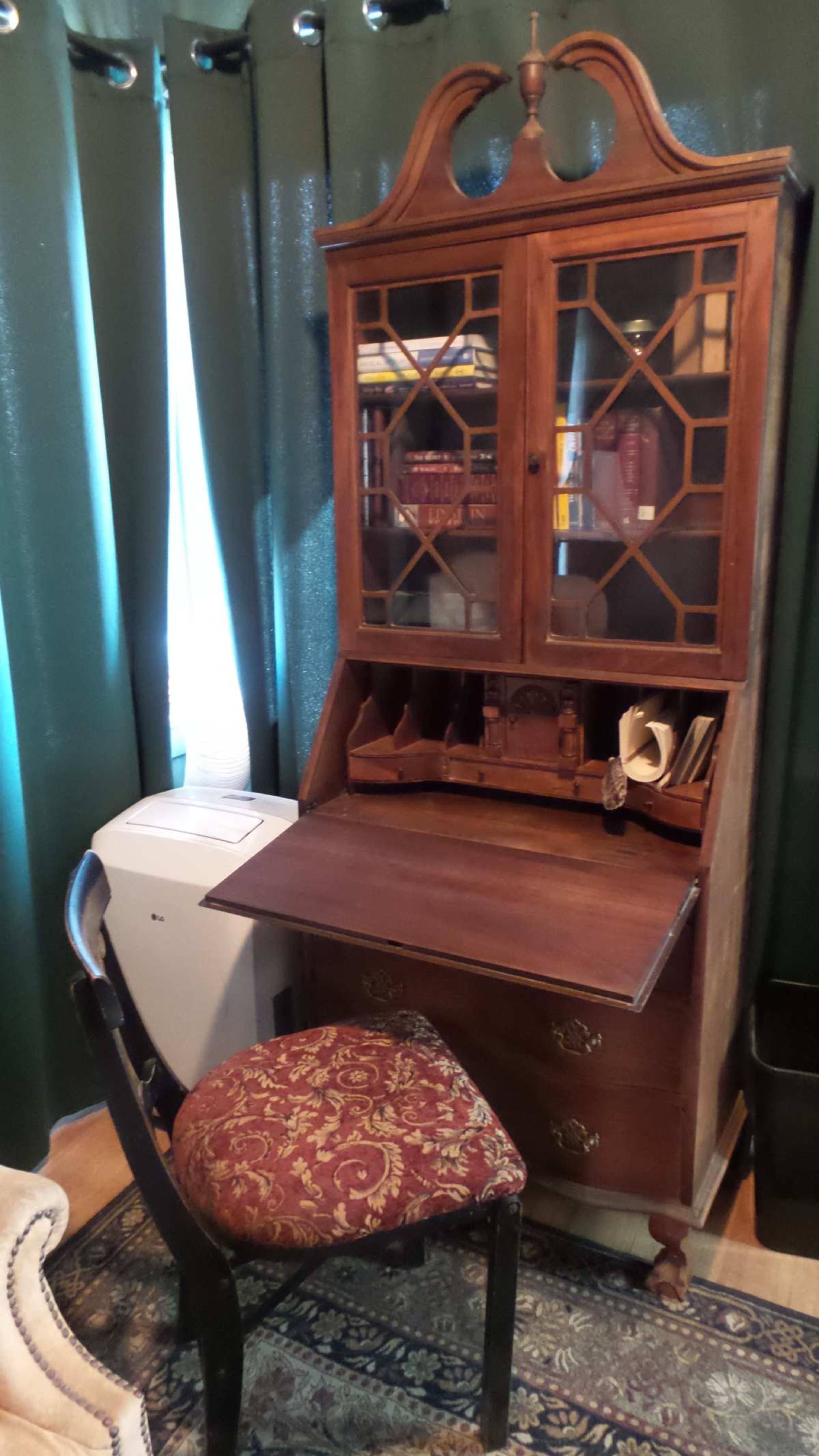 A wooden desk with a hutch, featuring multiple compartments, next to a black chair in a room with green curtains.