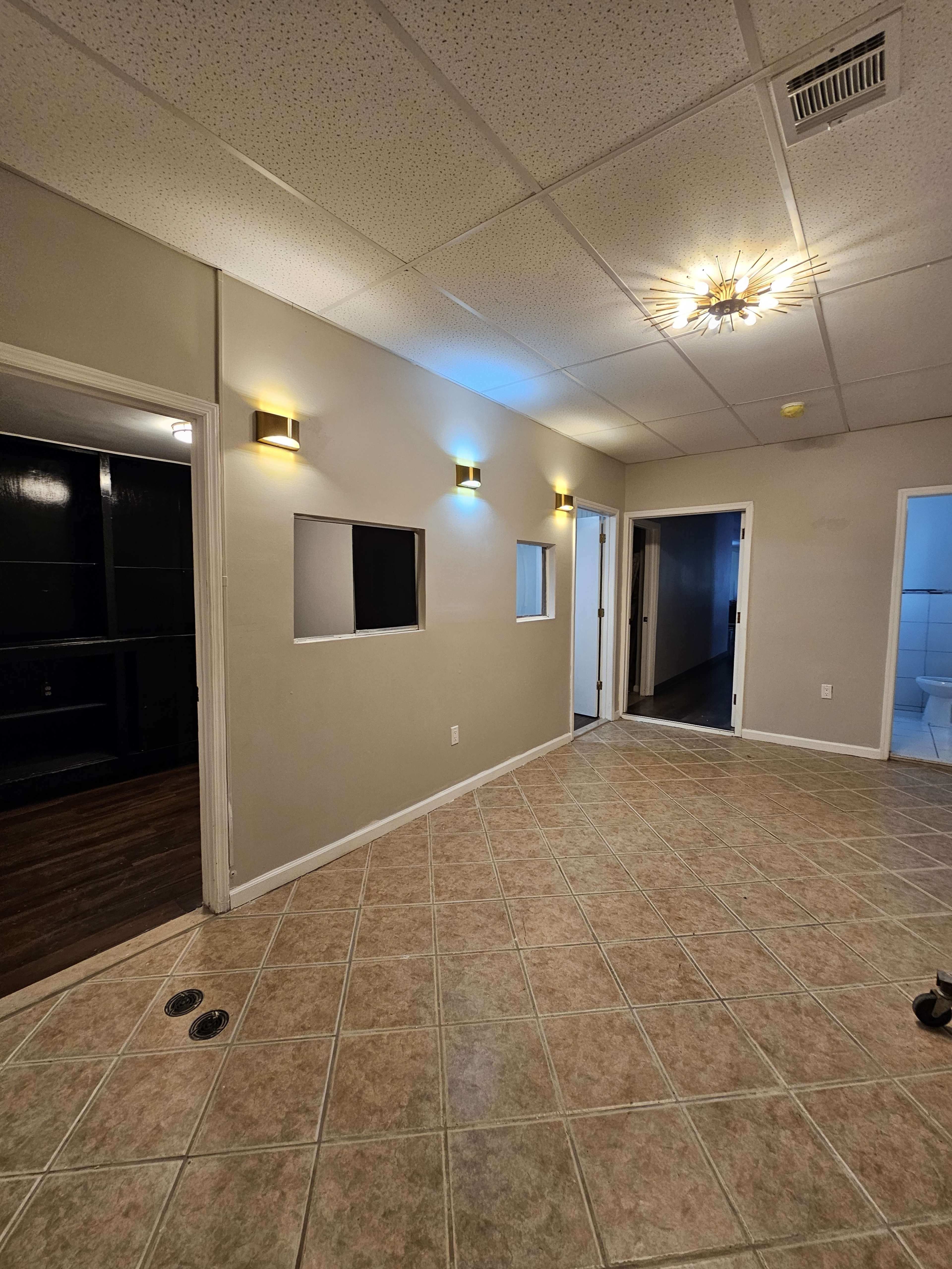 The image shows a room with tiled flooring, beige walls, two light fixtures, and two small window-like openings, leading to adjacent spaces.