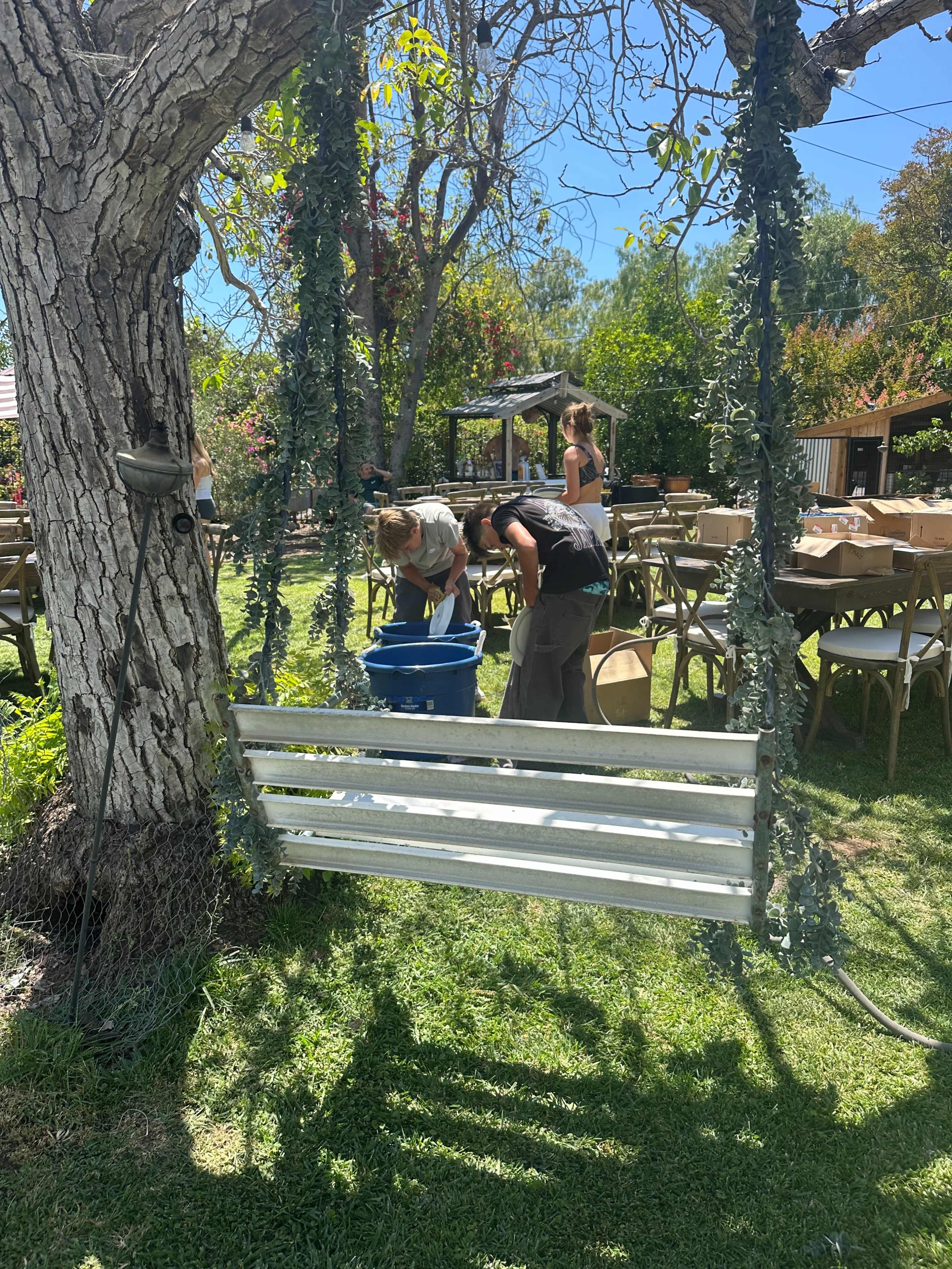 Two people work in front of a wooden swing beneath a tree, surrounded by tables and chairs set up in a garden.