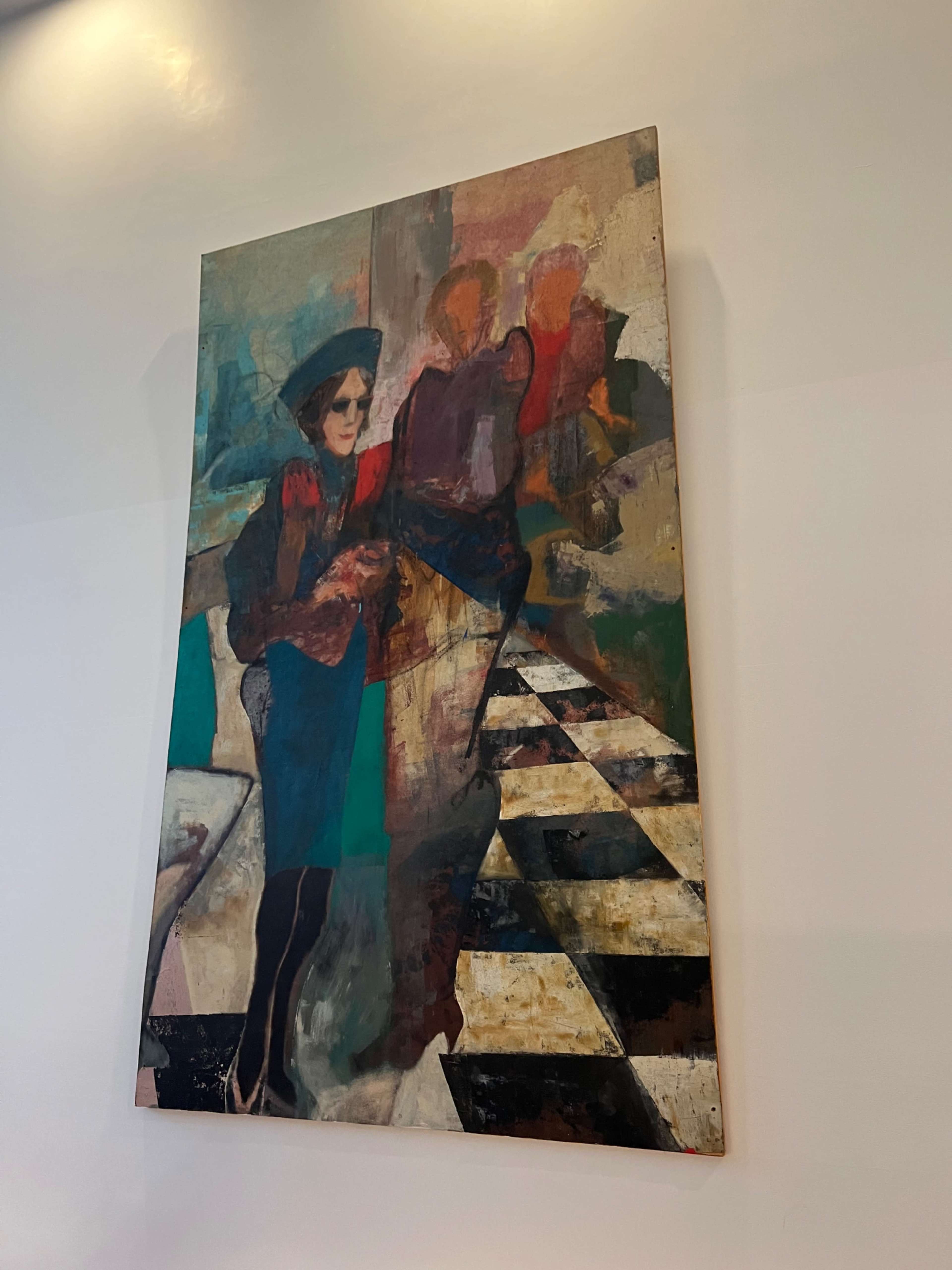A large painting hung on a wall, featuring a figure with a hat standing next to abstract, colorful shapes, with a black and white checkered pattern at the bottom.