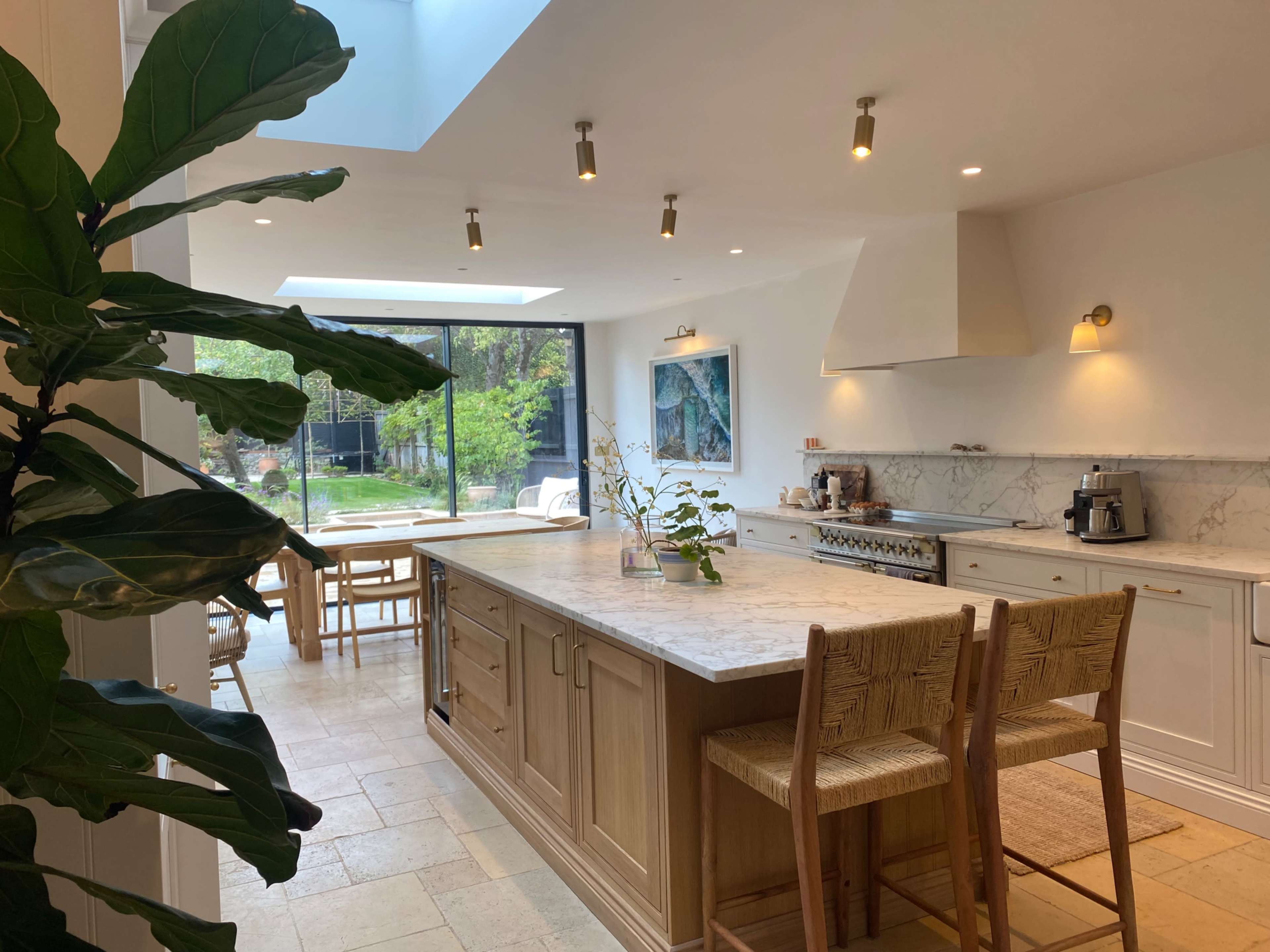 The image shows a modern kitchen with a large wooden island, marble countertops, and natural light streaming in from a skylight and large windows, overlooking a garden.