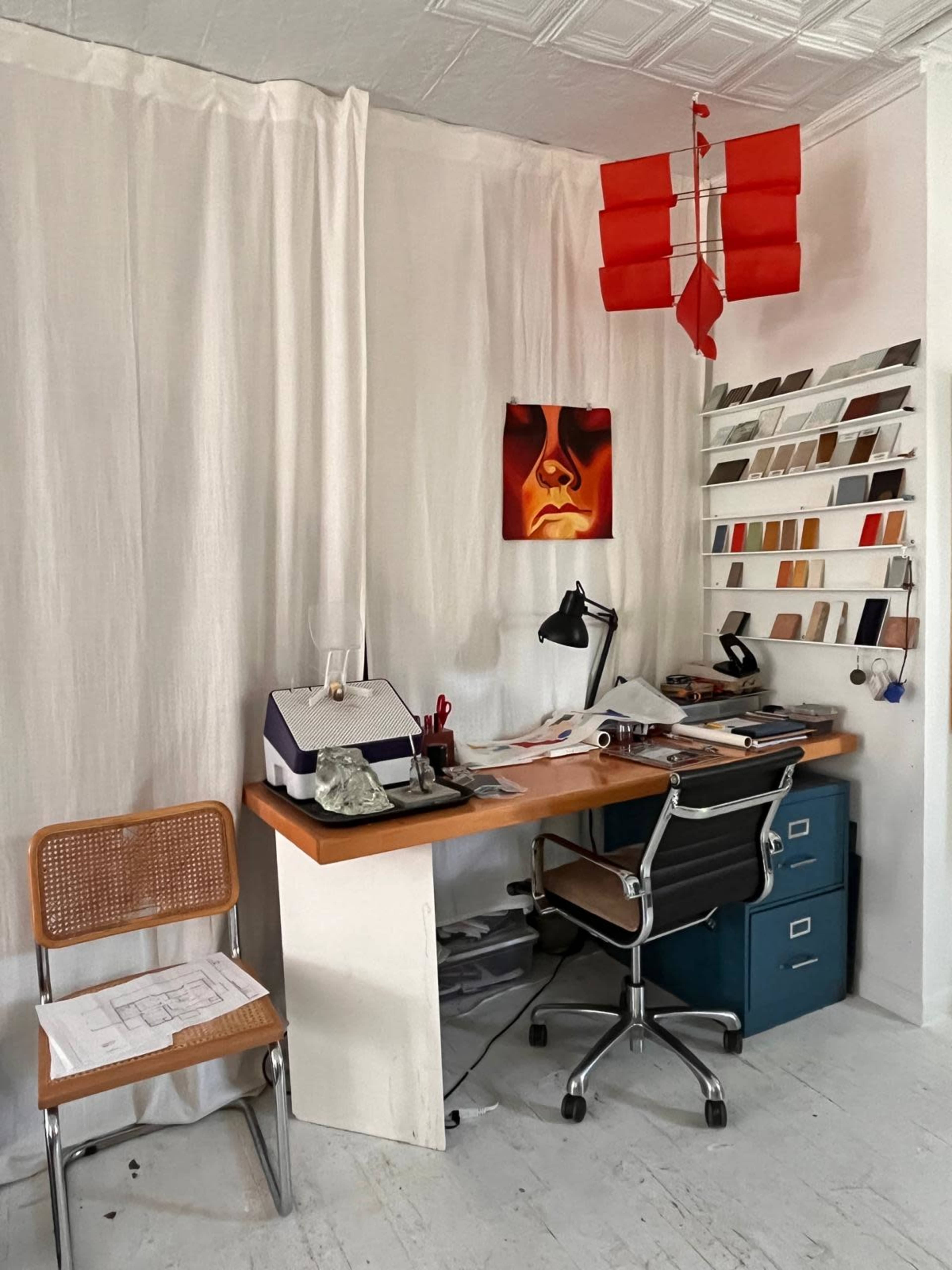 The image shows a minimalist workspace featuring a wooden desk with an office chair, lined shelves displaying color samples, and a wall adorned with a draped curtain and a framed artwork.