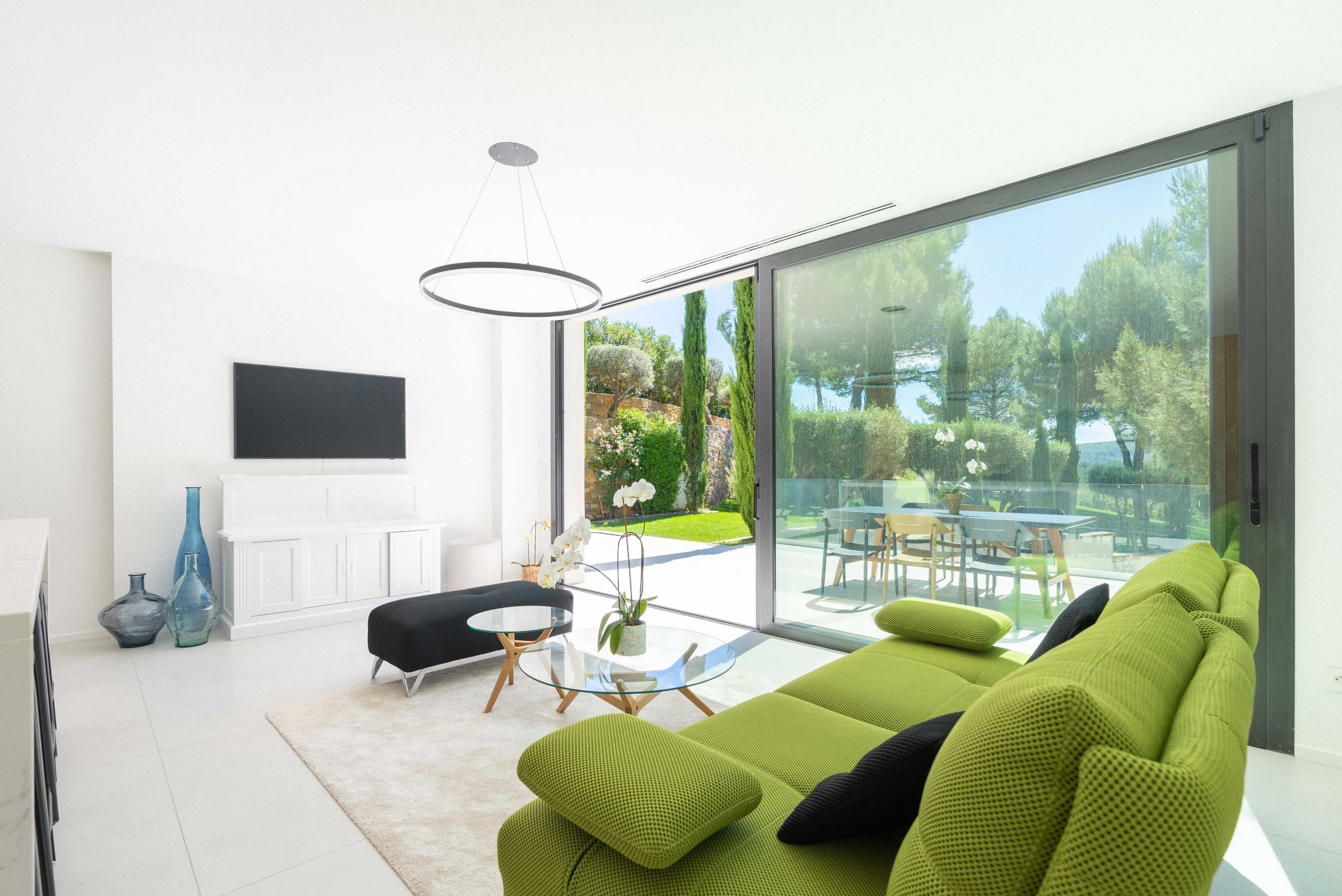 The image shows a modern living room with a green sofa, a glass coffee table, and large windows overlooking a garden area.