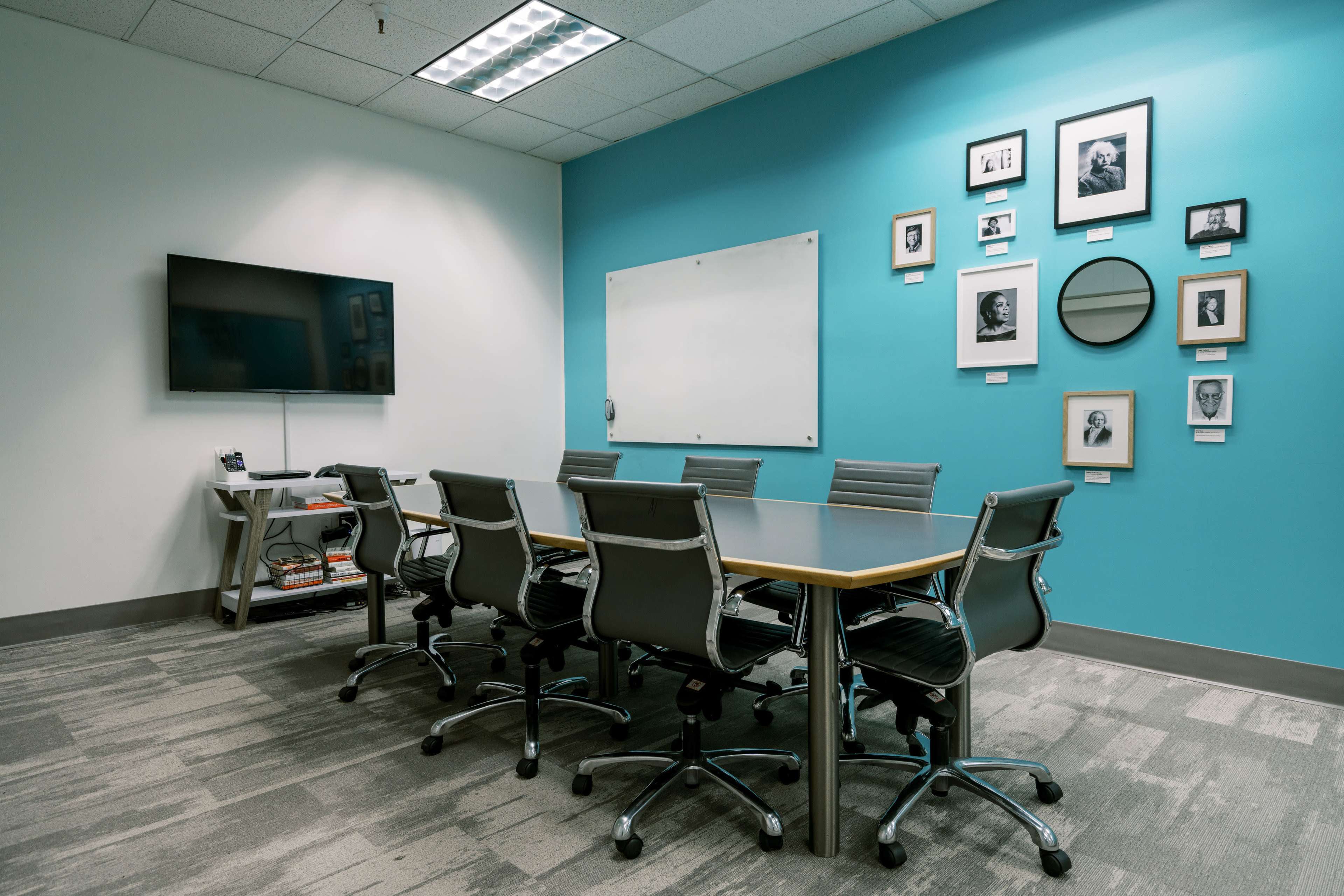 A conference room features a long table with several rolling chairs, a television on the wall, and a gallery of framed images on a bright blue wall.