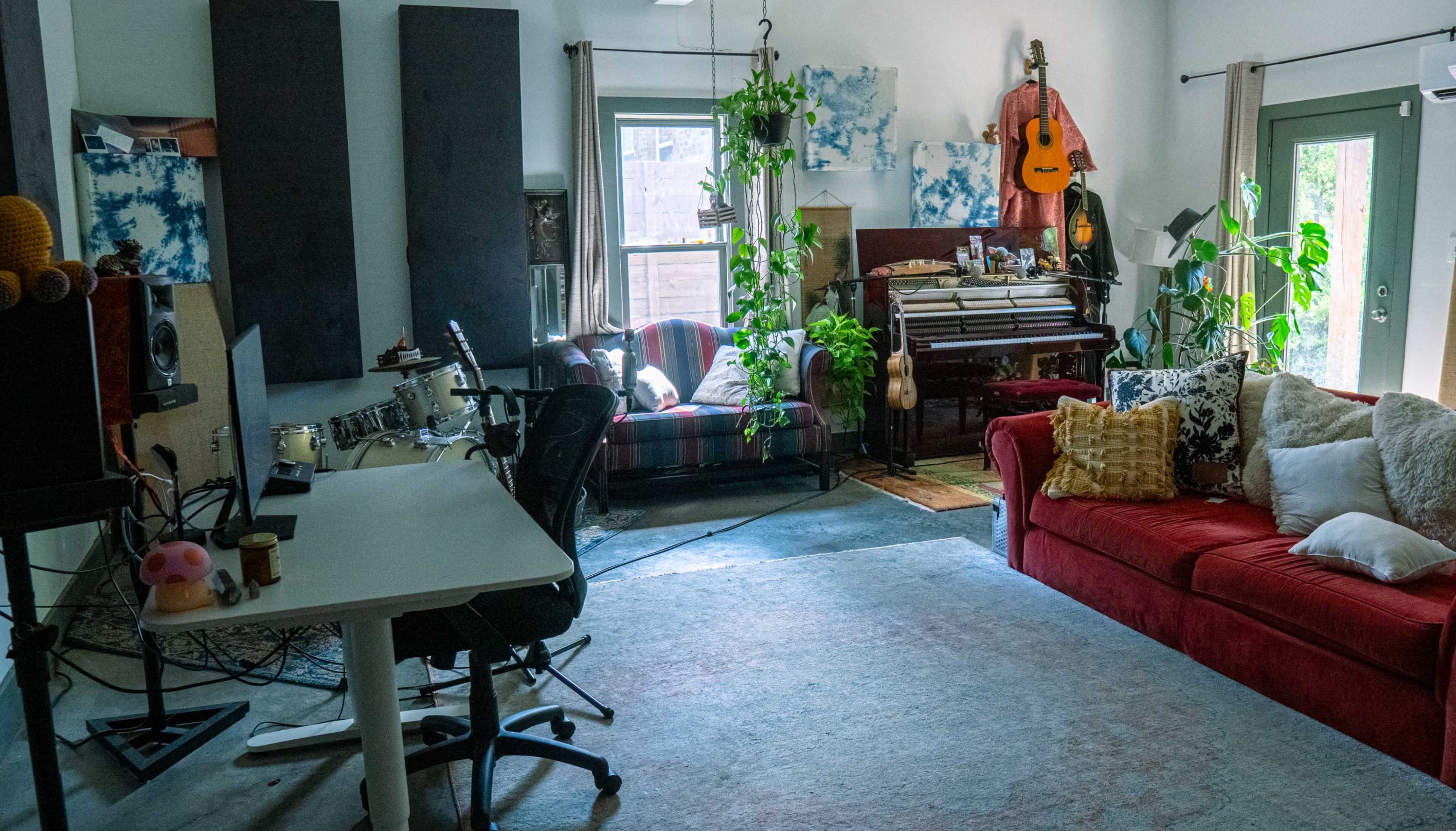 The room features a red couch, a wooden piano, a desk with musical equipment, and various plants along with colorful artwork on the walls.