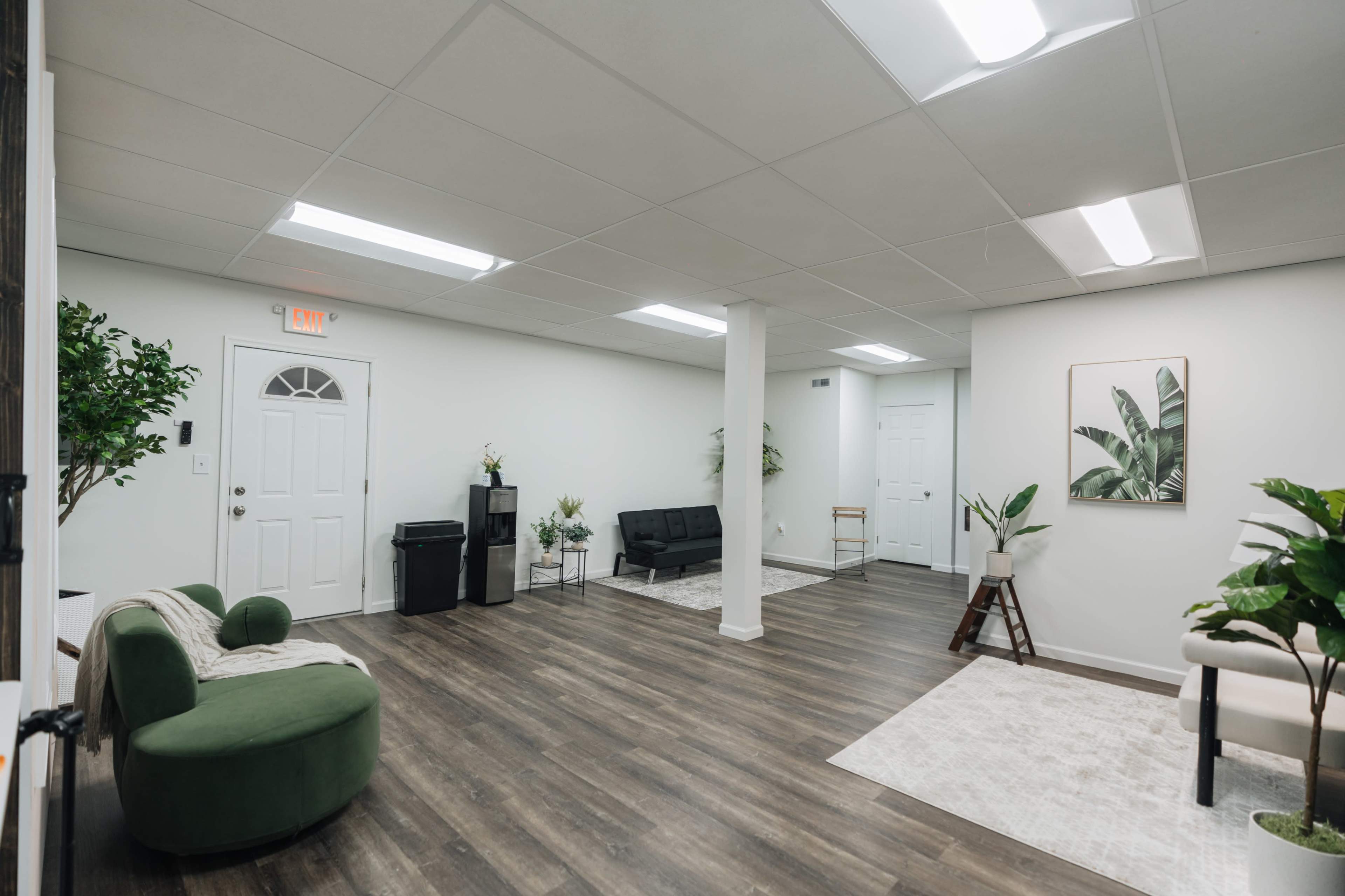 The image shows a spacious, well-lit interior with a mix of seating arrangements, plants, and a door leading to another room.
