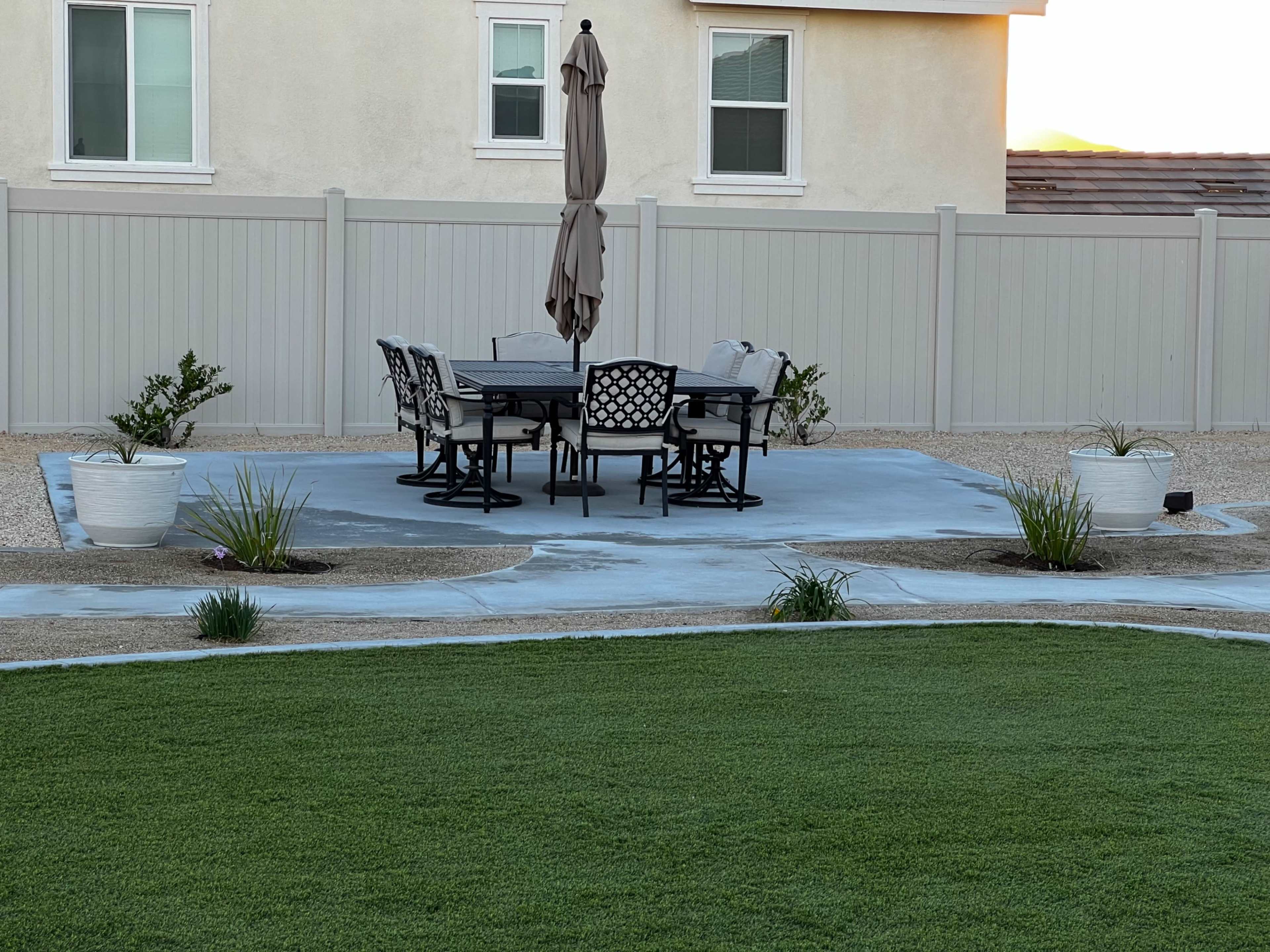 A circular concrete patio with a black and white dining set and an umbrella is situated in a landscaped yard surrounded by a low fence.