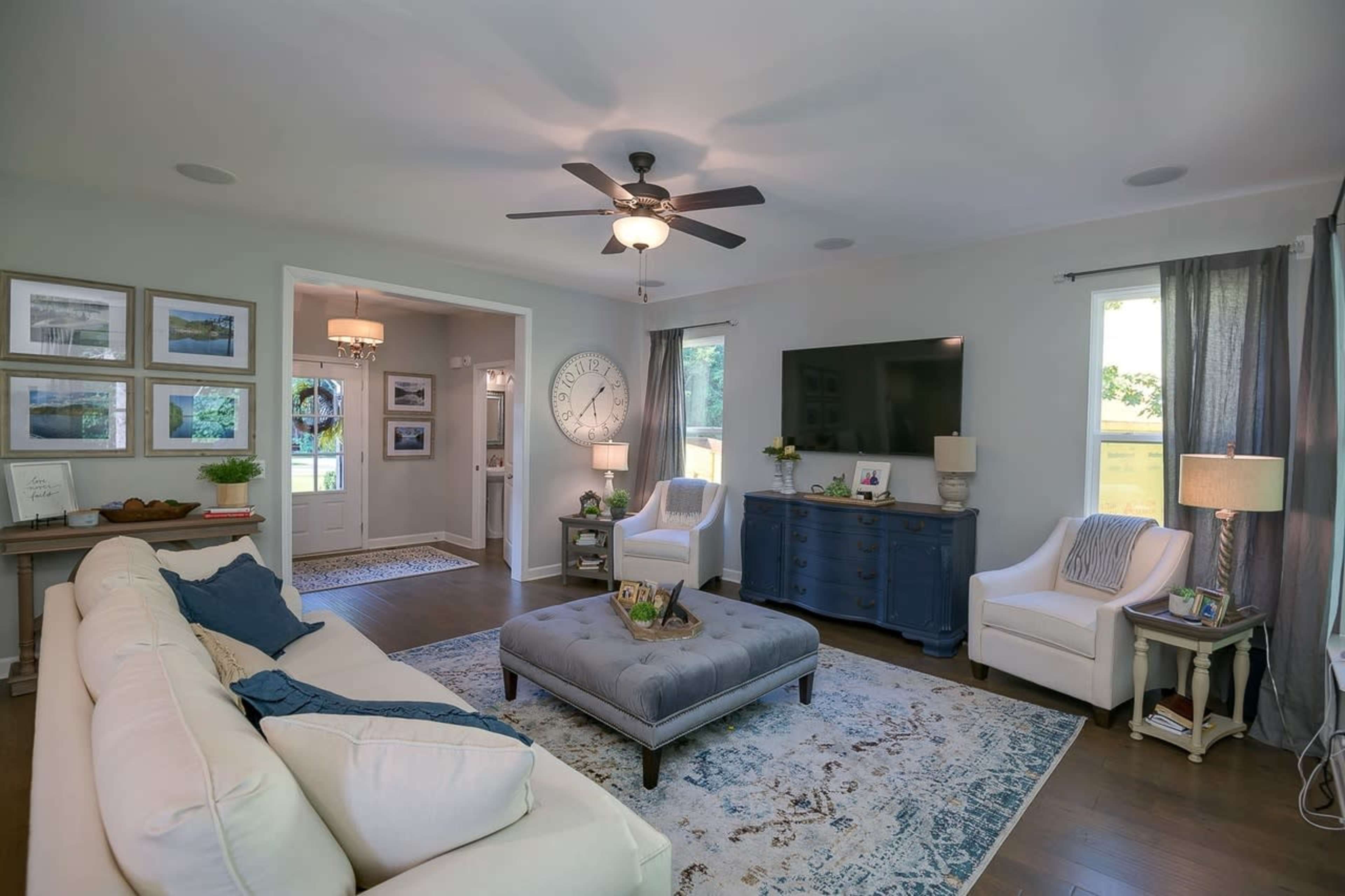 The image shows a modern living room with a beige sofa, blue ottoman, a dark wooden console table, and framed pictures on the walls, all illuminated by natural light from windows.