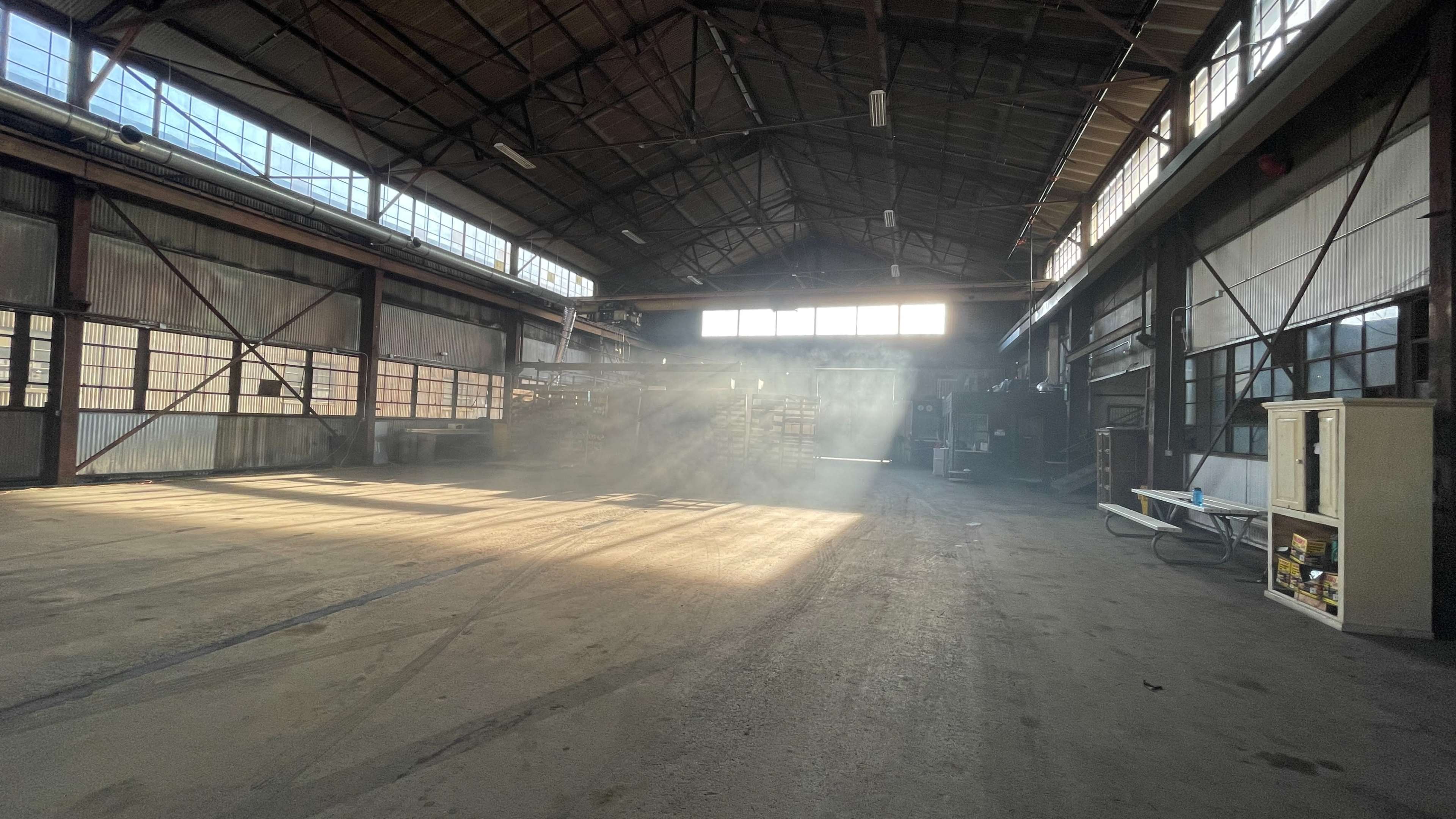 The image shows a spacious industrial warehouse with sunlight streaming through large windows, illuminating dusty air and empty floor space.