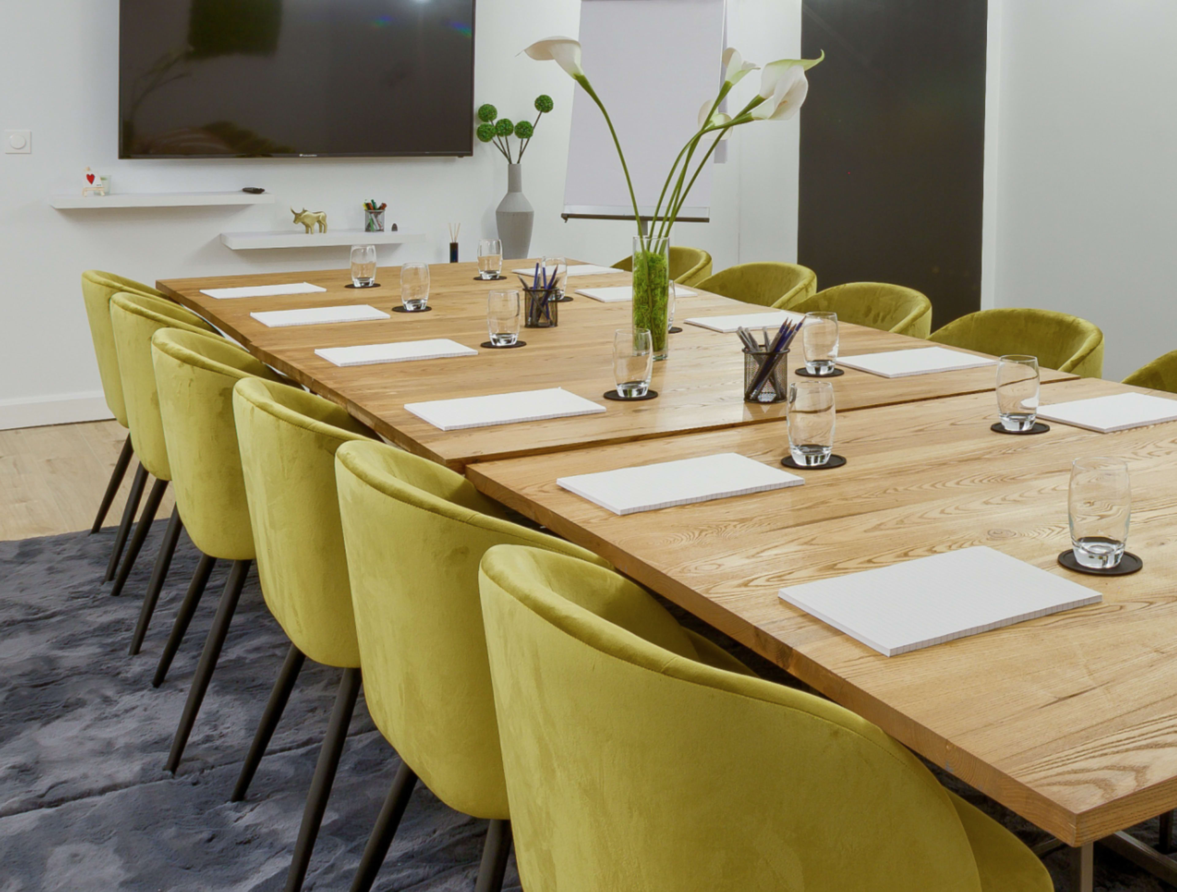 A long wooden conference table is set with white notepads and glasses of water, surrounded by green chairs in a modern meeting room.