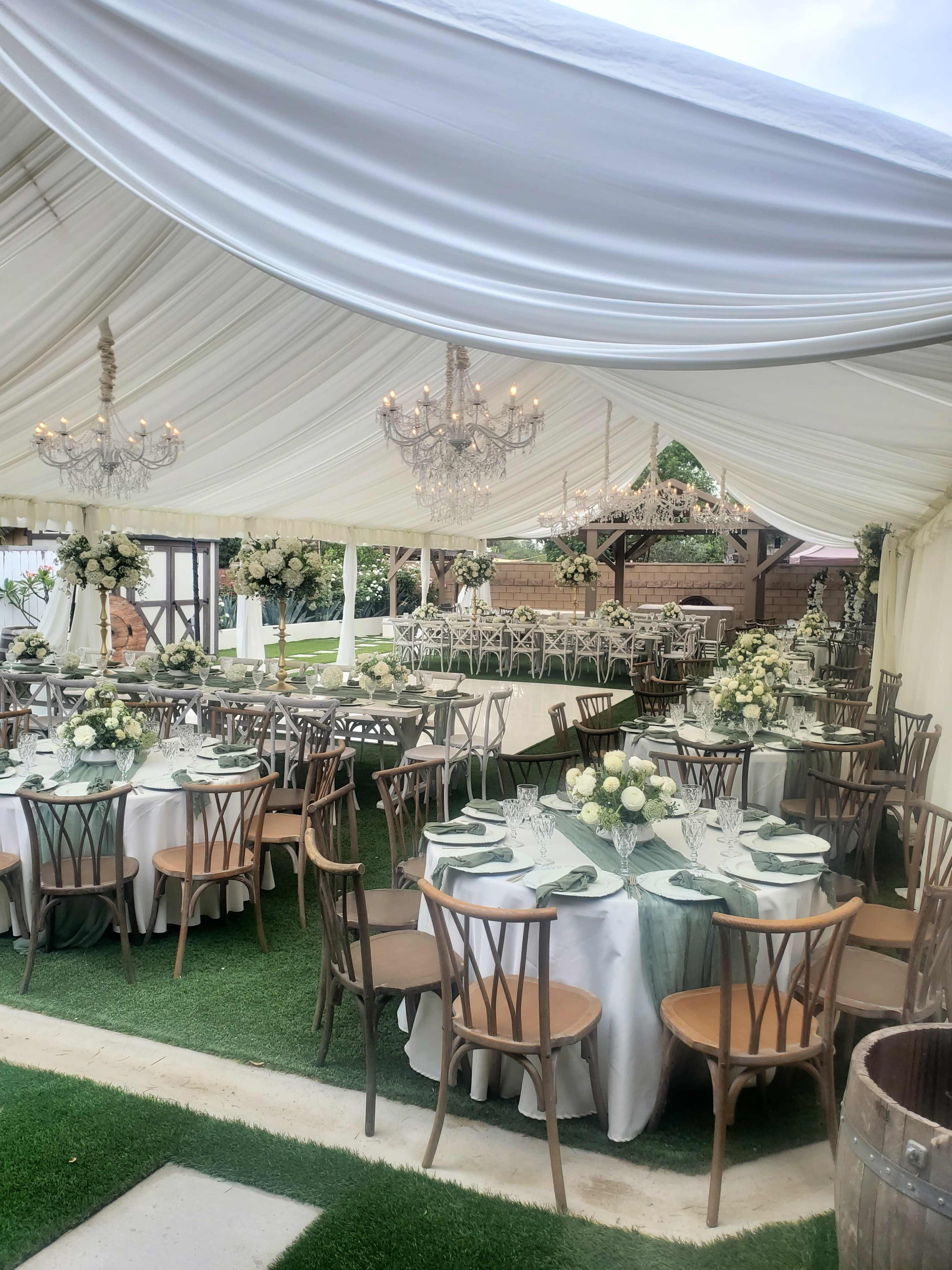 The image shows a beautifully arranged outdoor tented dining area with round tables set for a formal event, decorated with floral centerpieces and elegant chandeliers hanging from the ceiling.