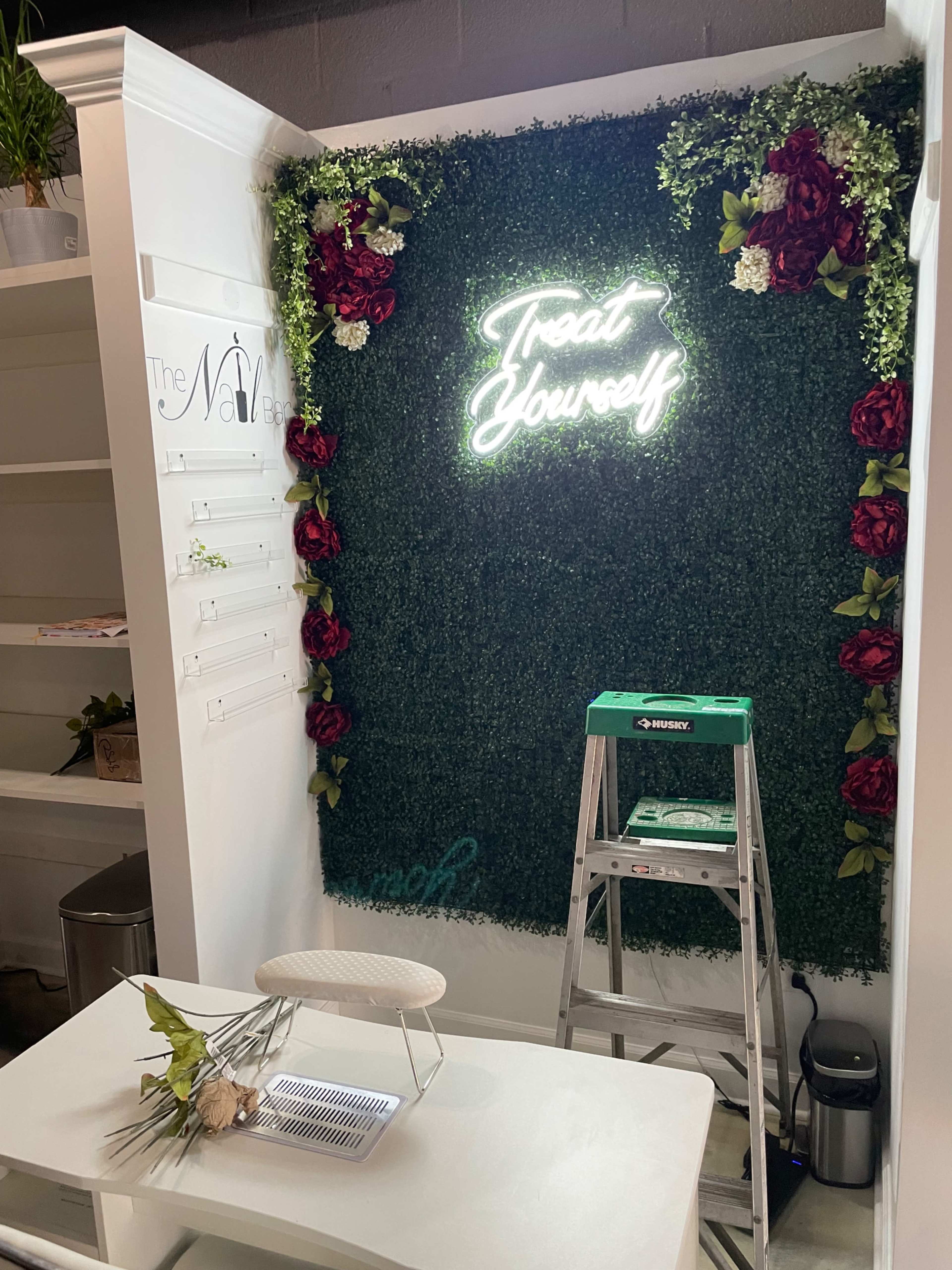 A decorative wall covered with greenery and flowers, featuring a neon sign that reads "Treat Yourself," alongside a small table and a step ladder.