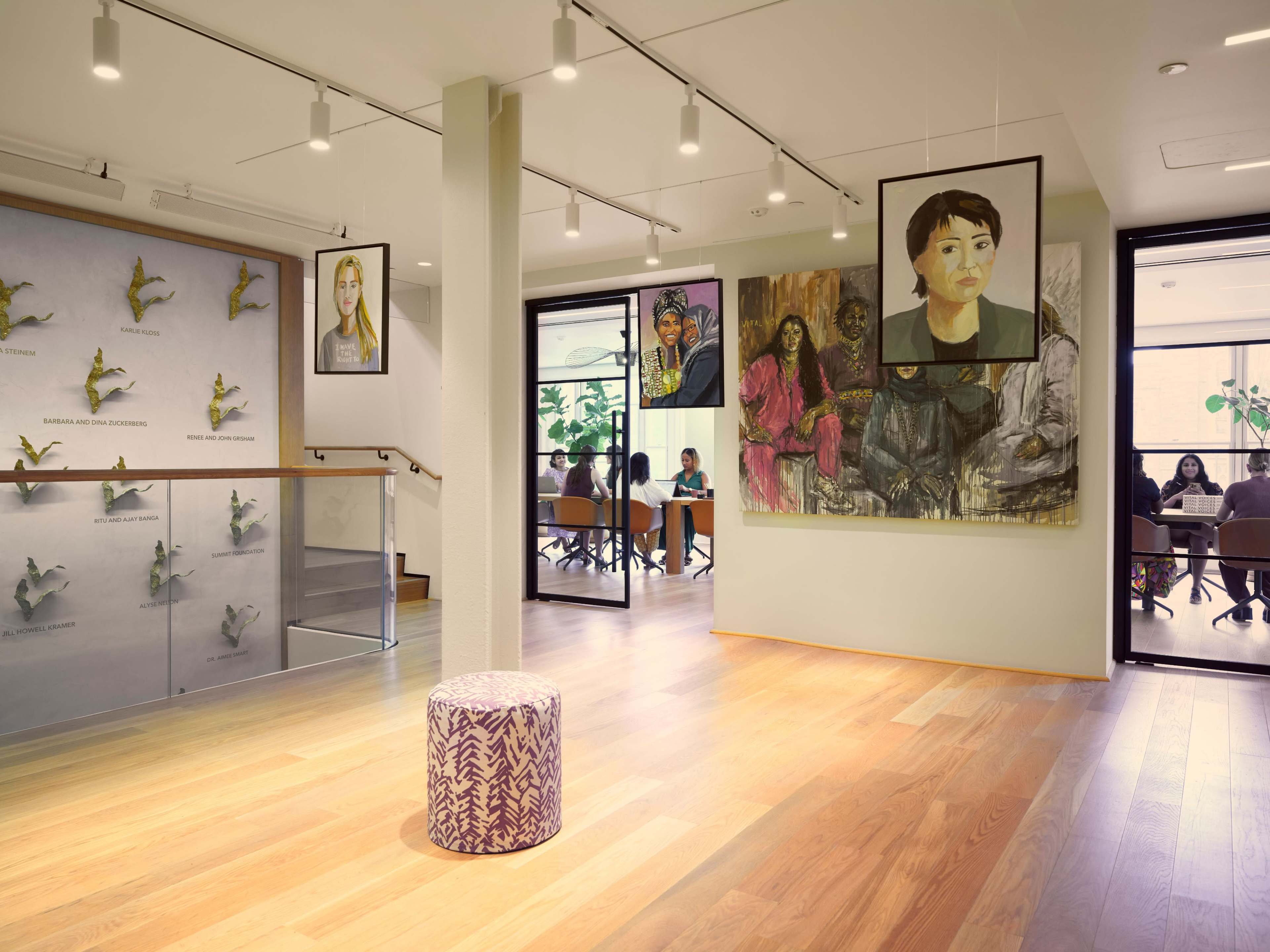 The image shows a modern, brightly lit interior space featuring several paintings on the walls and a small round stool in the center.