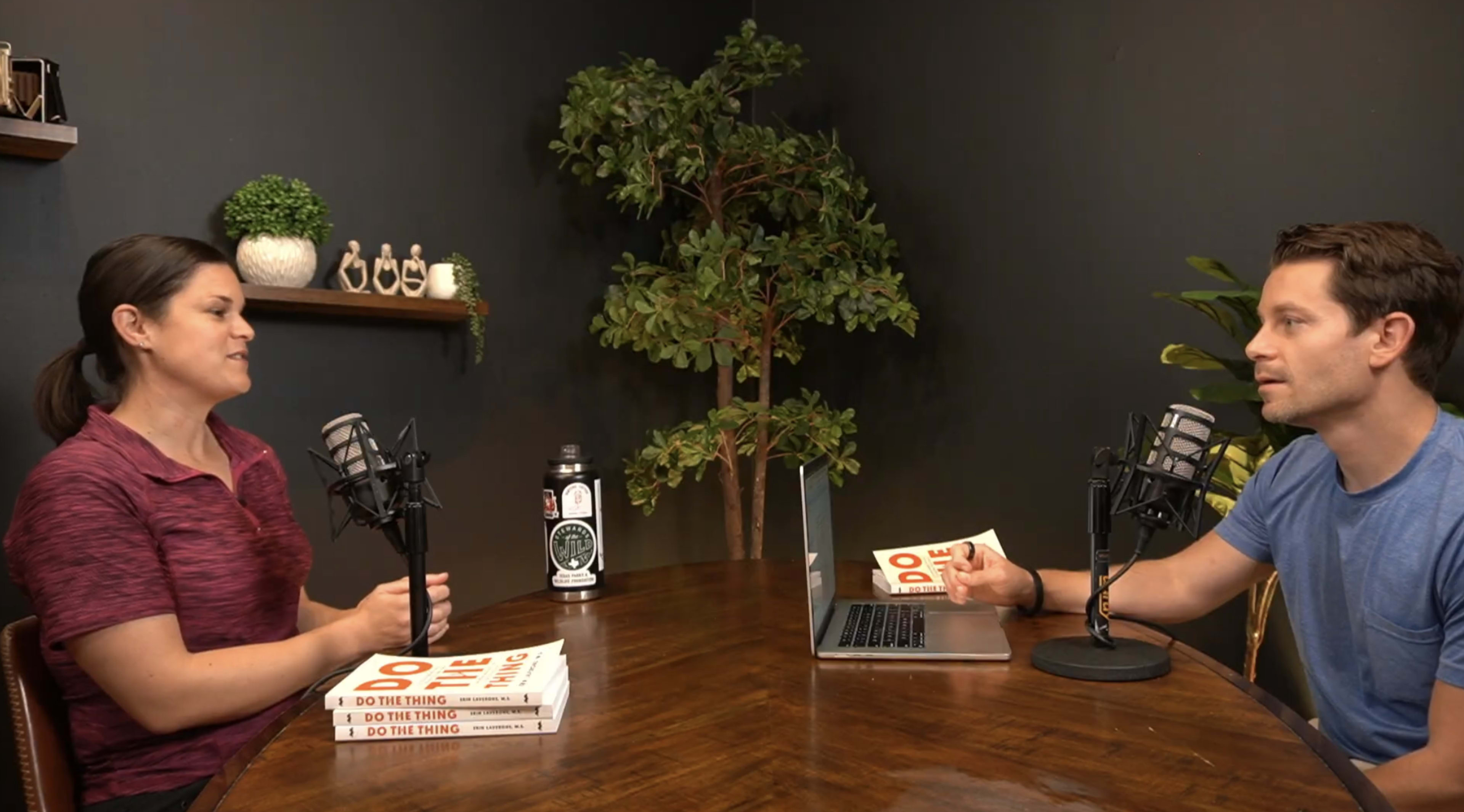 Two individuals are sitting at a round table with microphones, engaged in a conversation, while books are stacked on the table and a laptop is open in front of them.