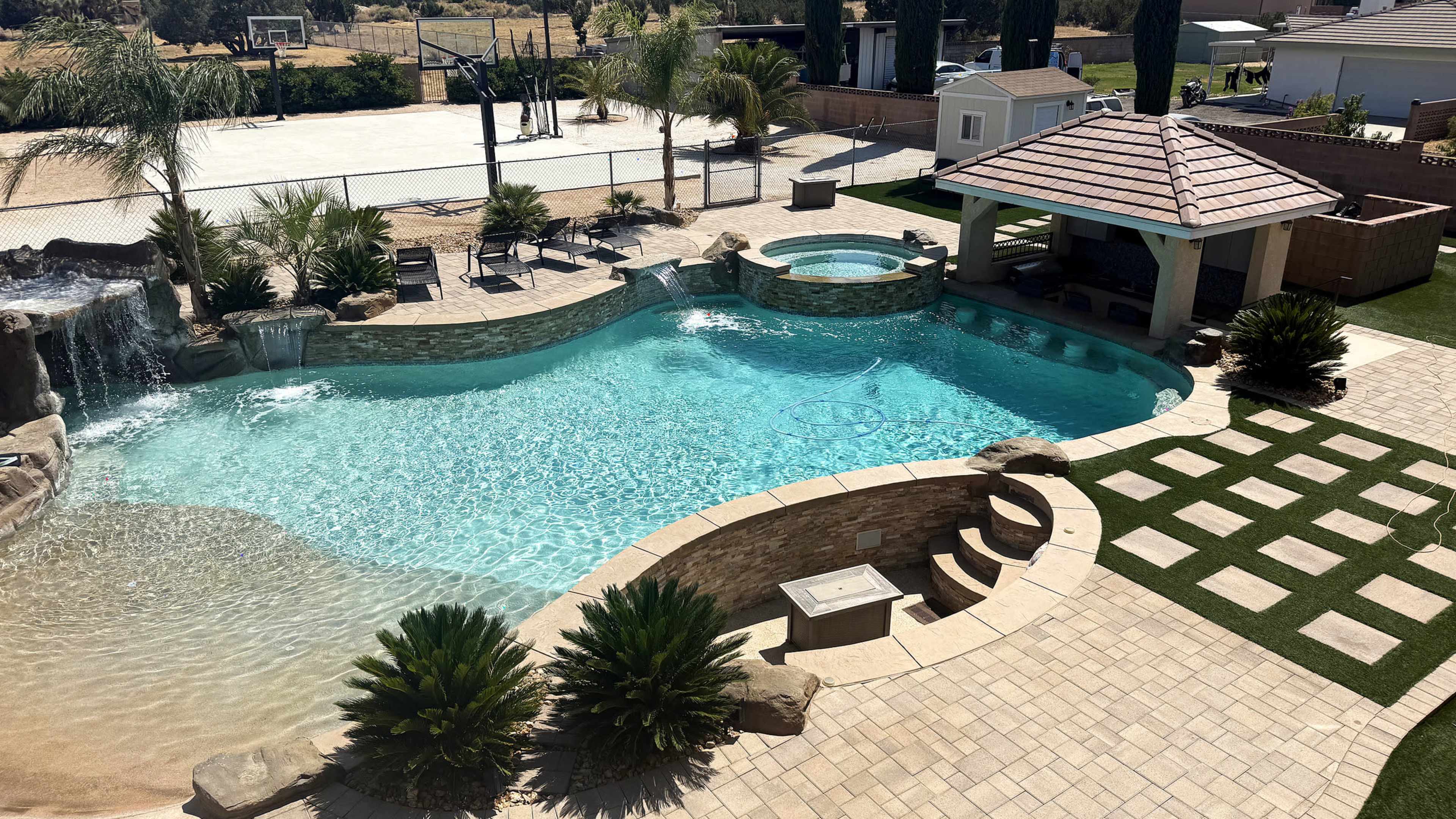 The image shows a large swimming pool with a waterfall feature, surrounded by palm trees and a patio area, with a basketball court visible in the background.