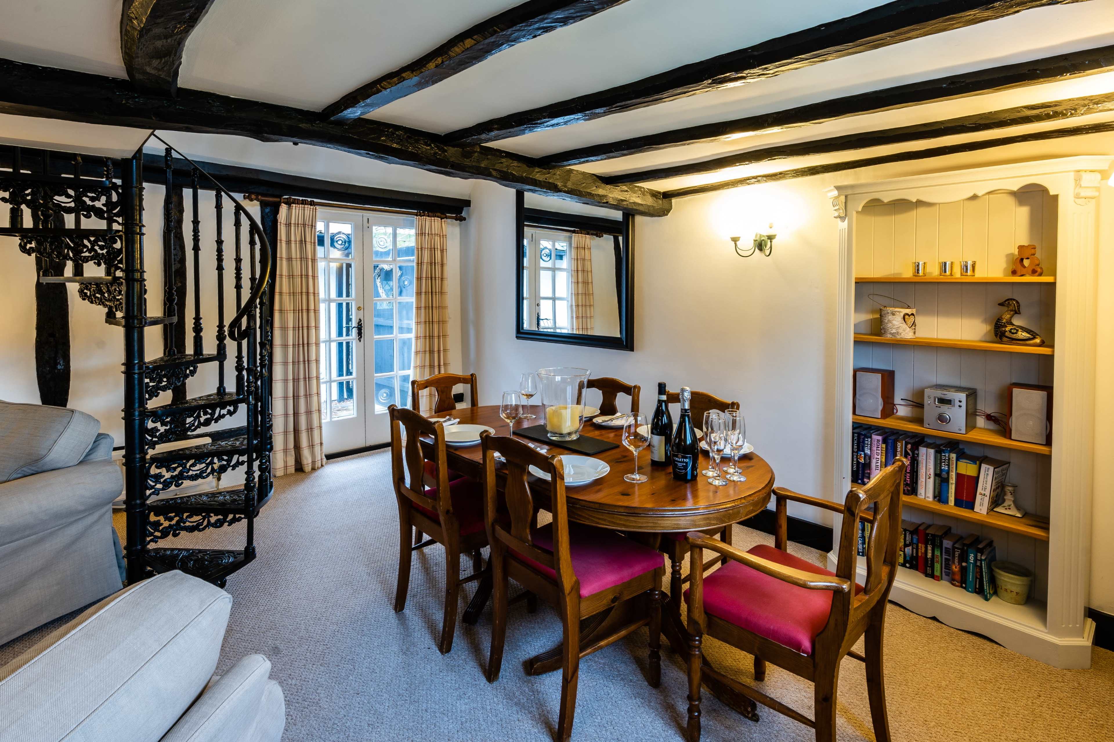 A dining area with a round wooden table set for a meal, surrounded by chairs, with a spiral staircase and bookshelves in the background.