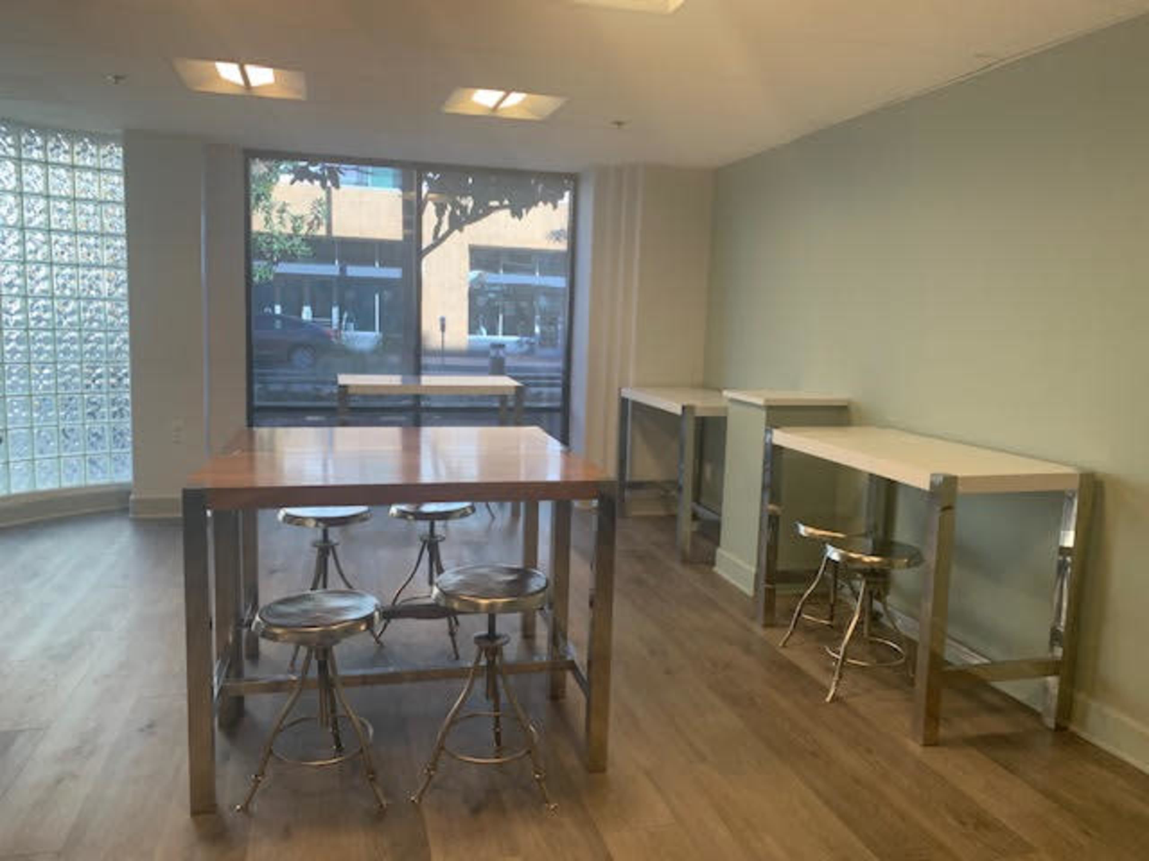 A room with several tall, rectangular tables and metal stools, featuring large windows that let in natural light.