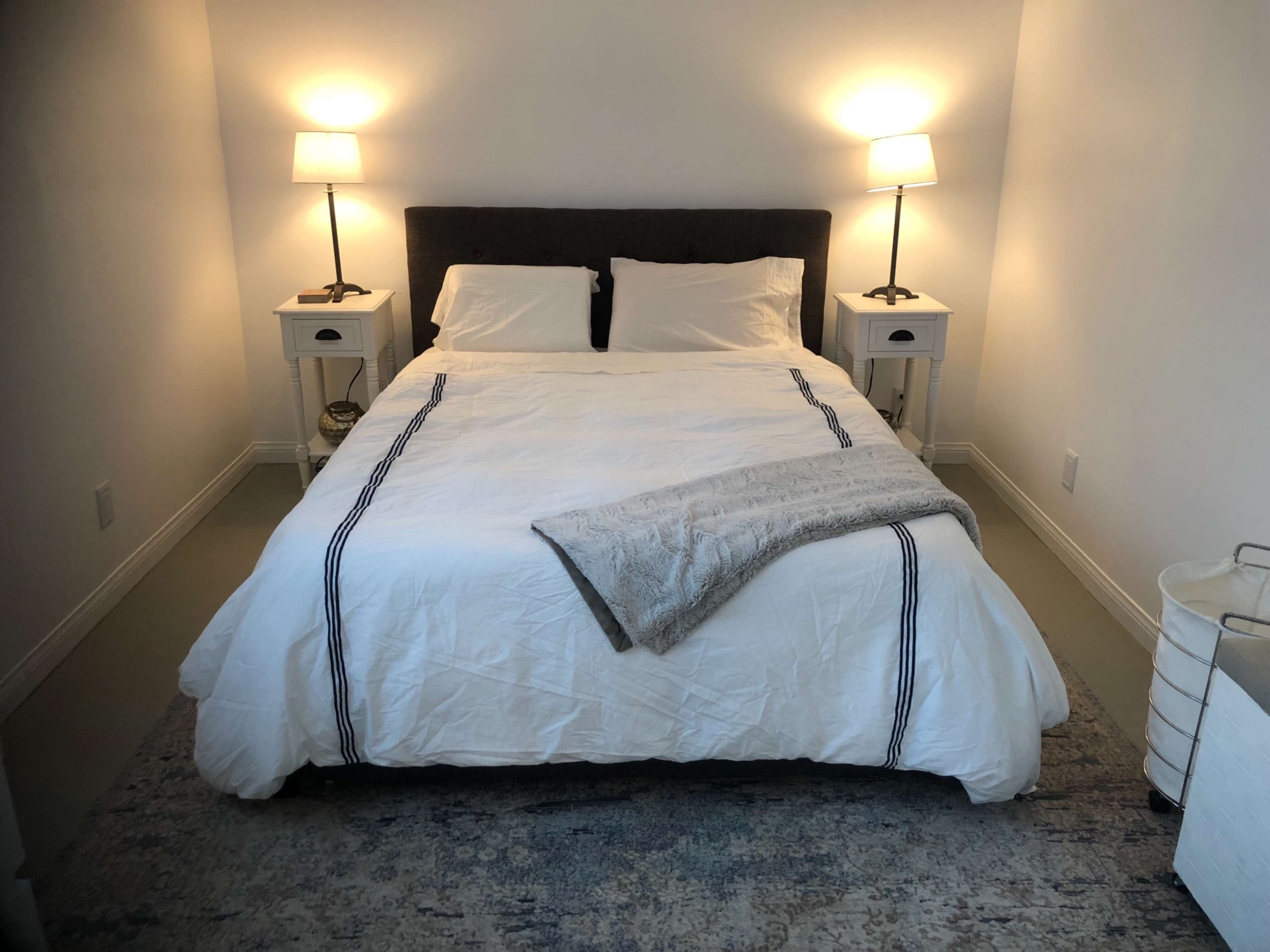 A neatly made bed with white linens and a gray throw is placed between two nightstands with lamps in a softly lit room.