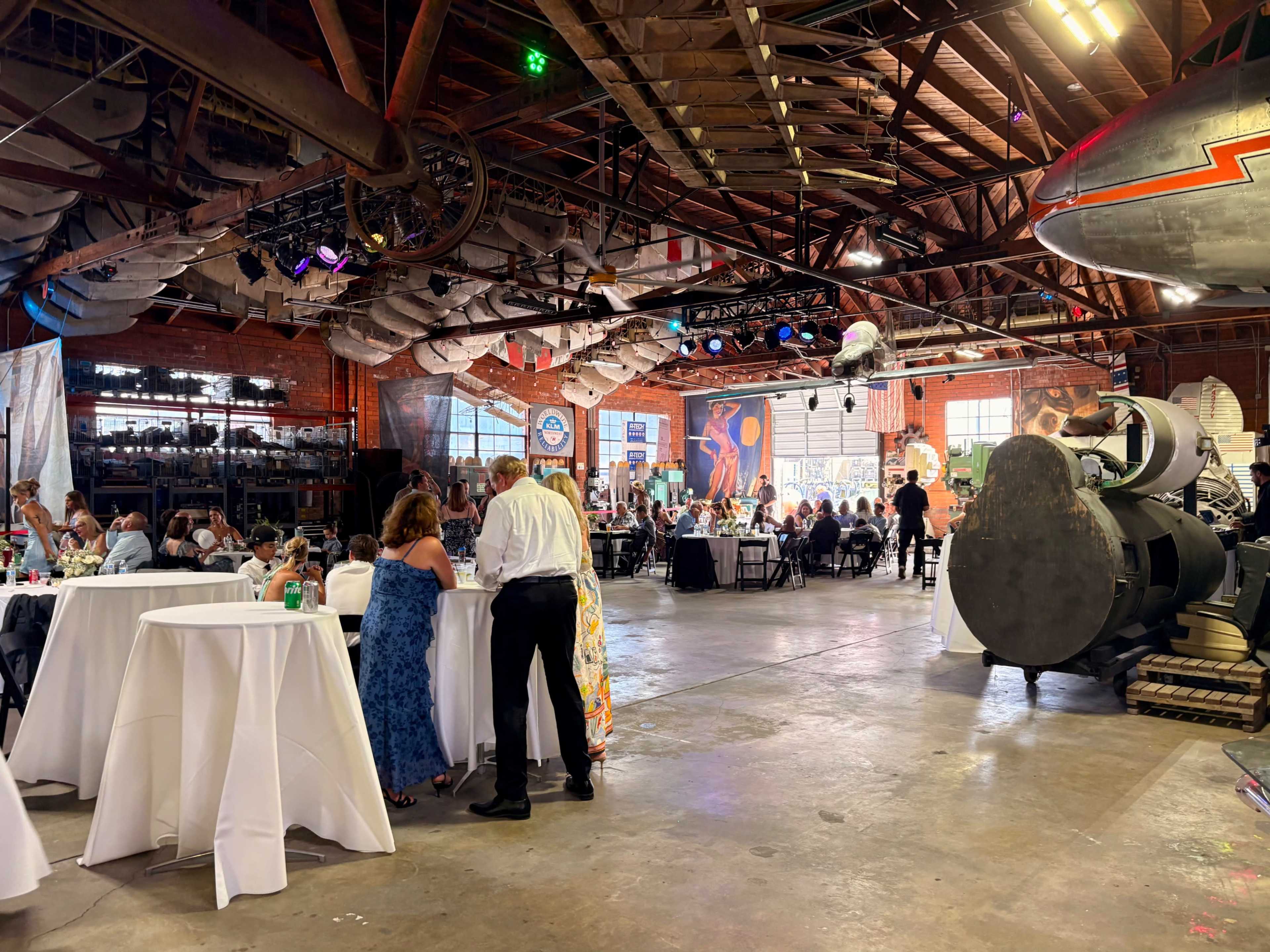 A spacious event venue with round tables and guests mingling, featuring large industrial elements and colorful decorations in the background.
