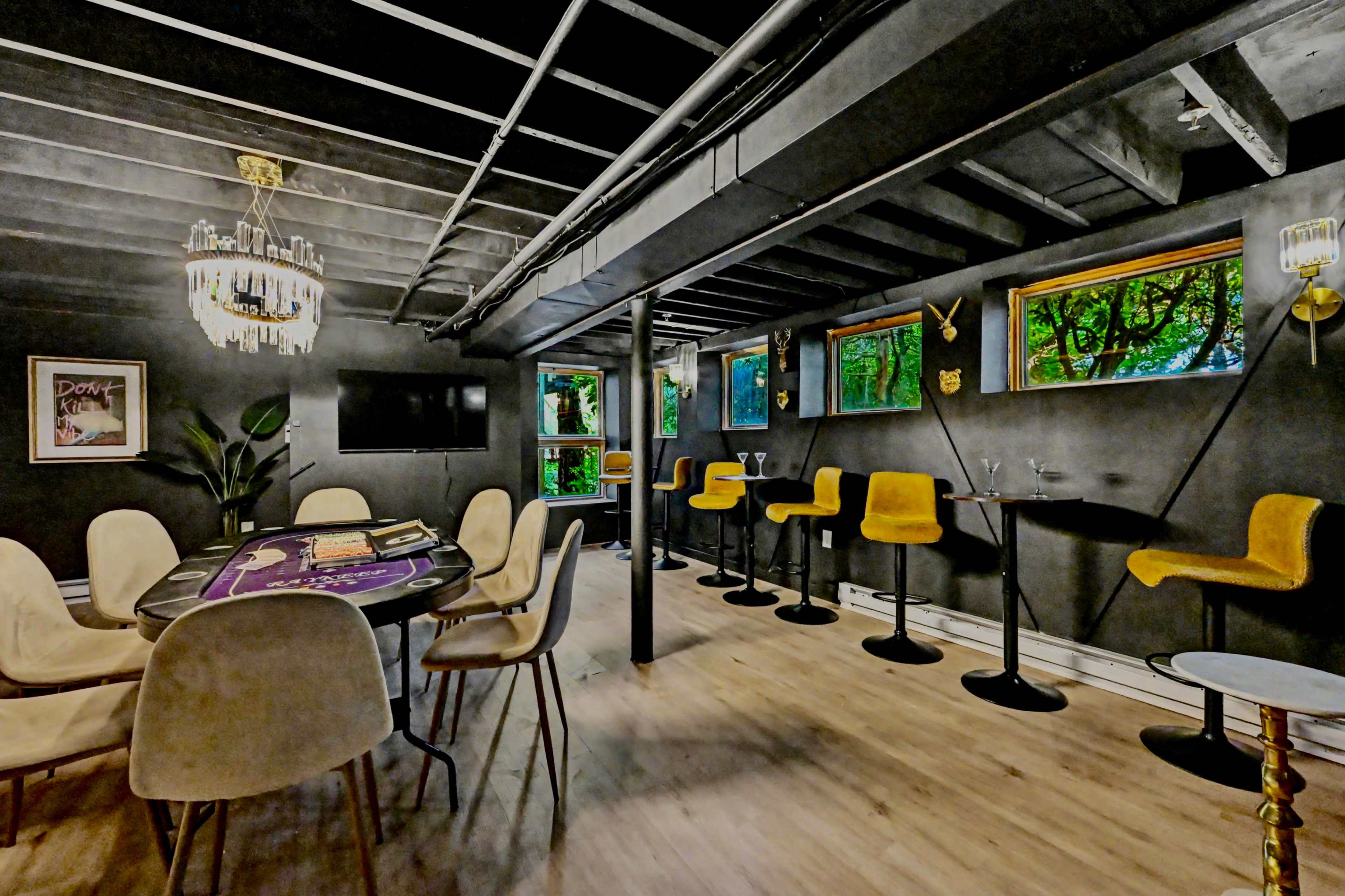 The image shows a modern lounge area featuring a poker table, seating at high-top tables, and large windows offering views of greenery outside.