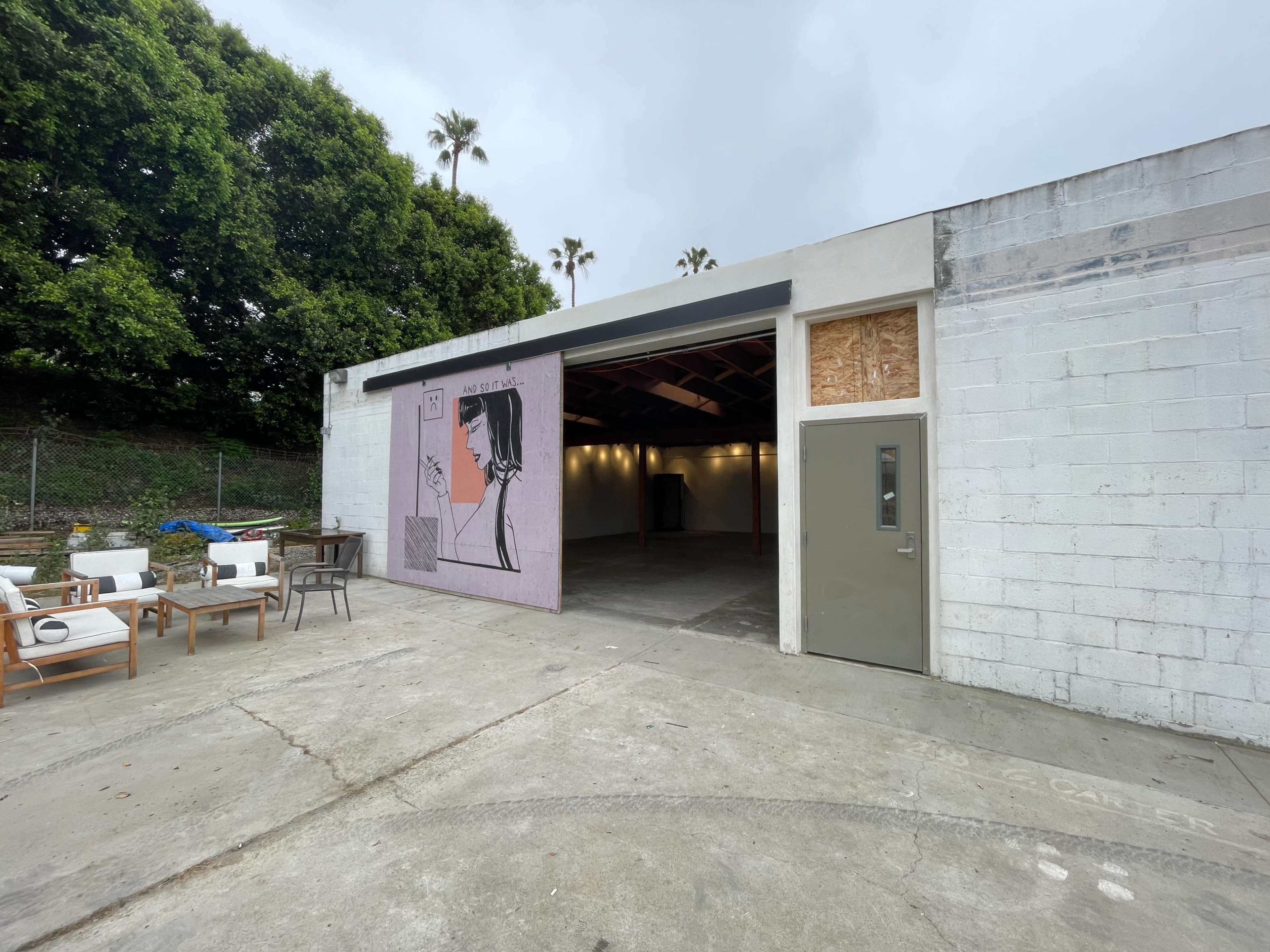 A large white building with partially open doors reveals an empty interior space, while a mural of a woman is visible on the exterior wall.