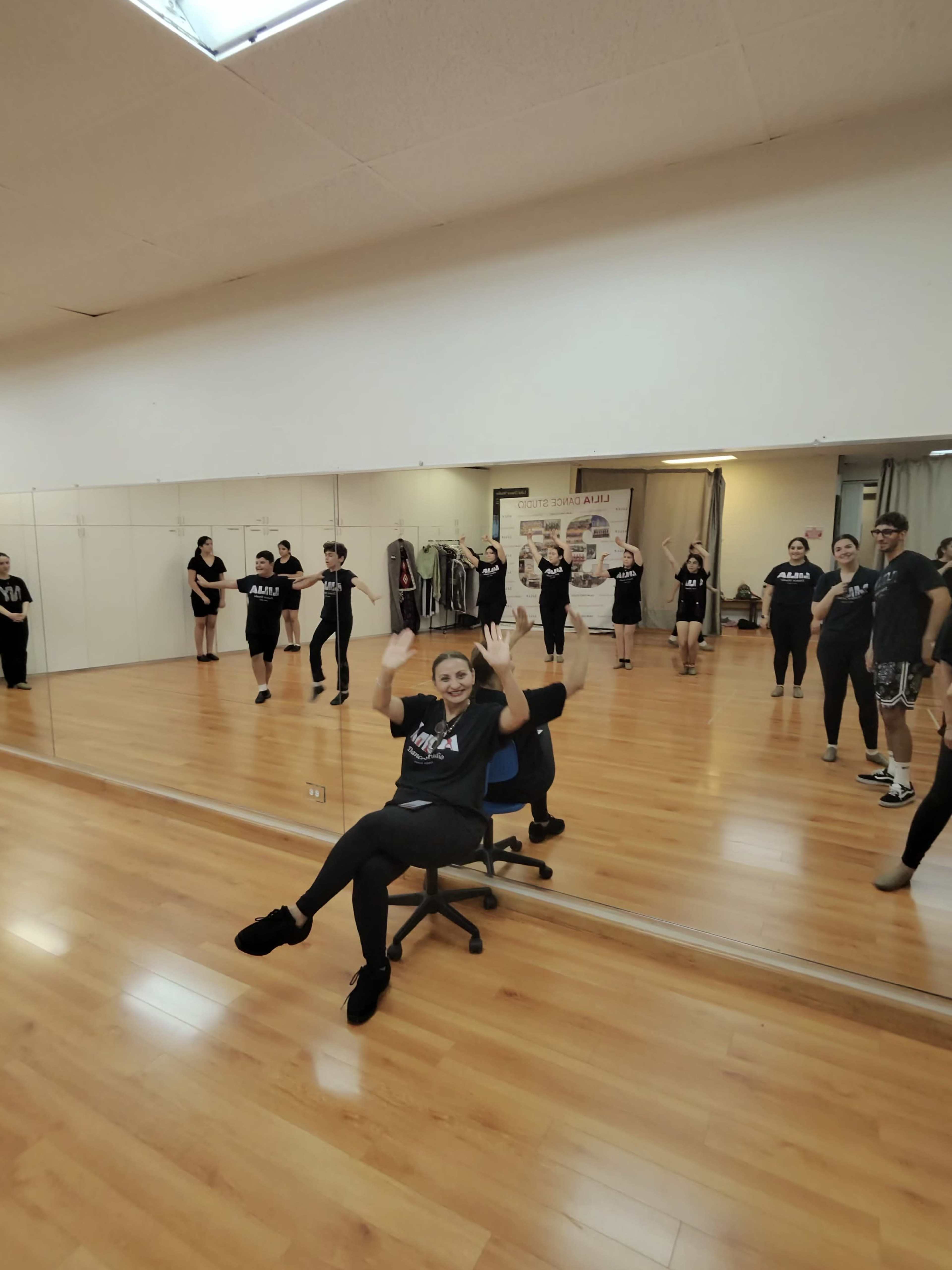 A group of dancers is practicing in a mirrored studio, with one dancer seated on a chair in the foreground.
