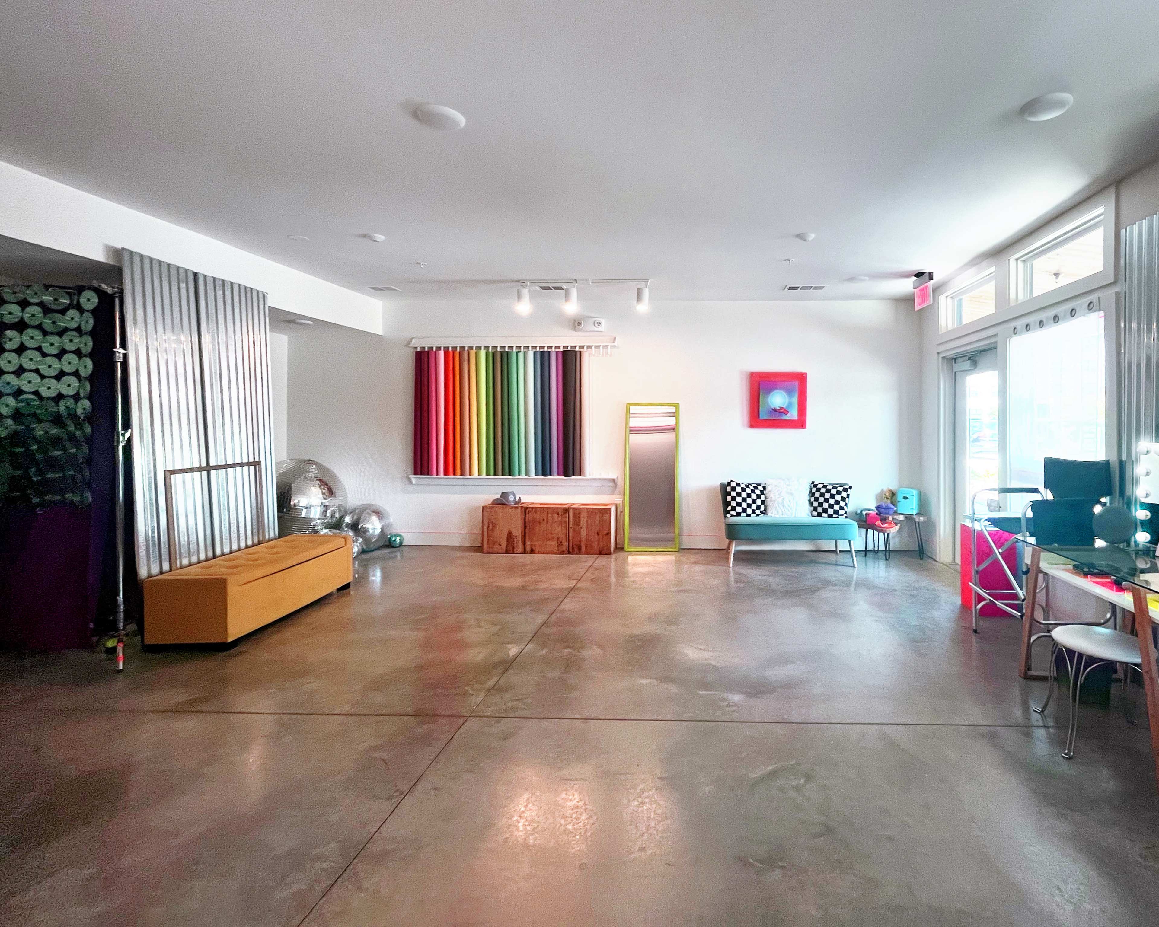 The image shows a spacious, brightly lit room with colorful artwork, a seating area, and polished concrete floors.